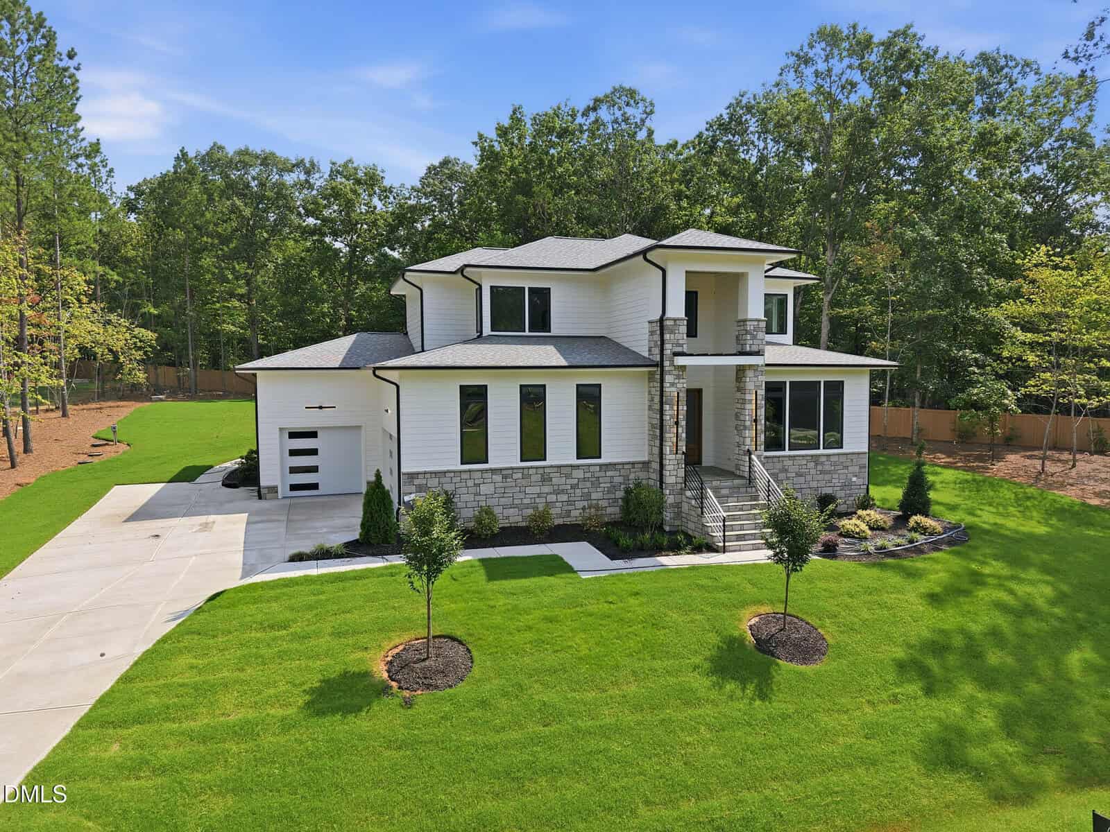 A two-story modern house with white siding and stone accents, large windows, a two-car garage, a manicured lawn, landscaping, a concrete driveway, and surrounded by trees under a clear blue sky.