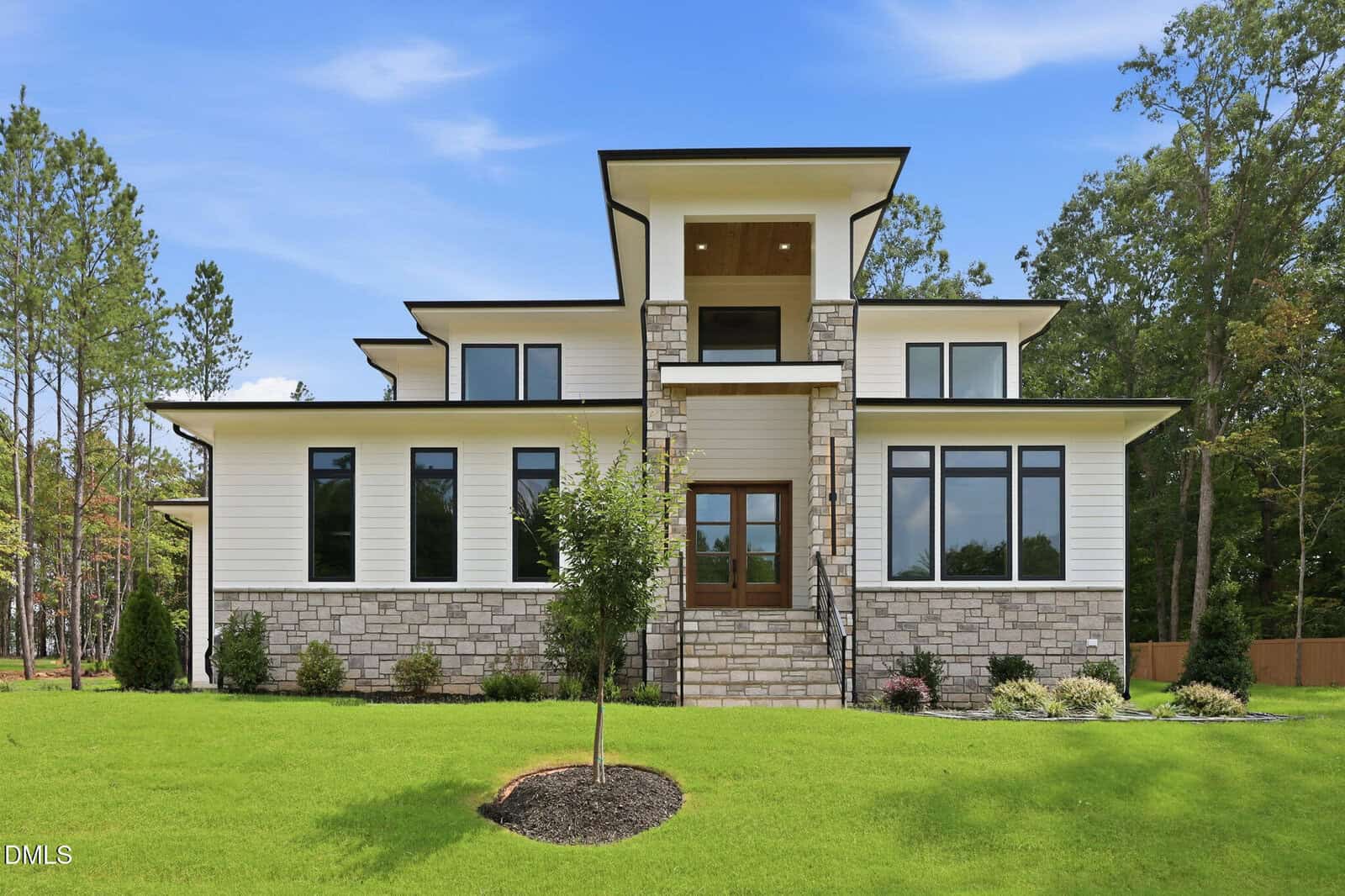 Modern two-story house at 20 Cherry Bark Drive in Youngsville, featuring a stone and white siding exterior, large windows, centered wood front door, sloped roof, inviting front steps, landscaped yard, and trees in the background under a blue sky.