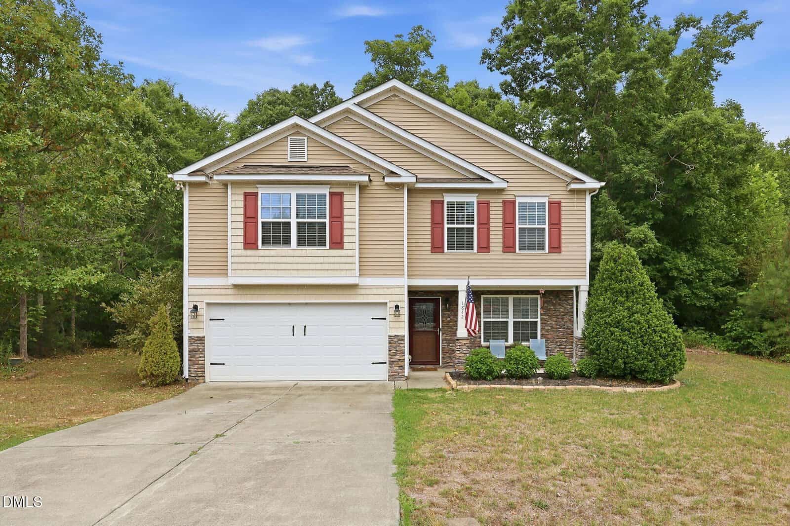 Two-story suburban house with tan siding, red shutters, a double garage, American flag by the entrance, and a driveway. The house is surrounded by trees and has a small front lawn.