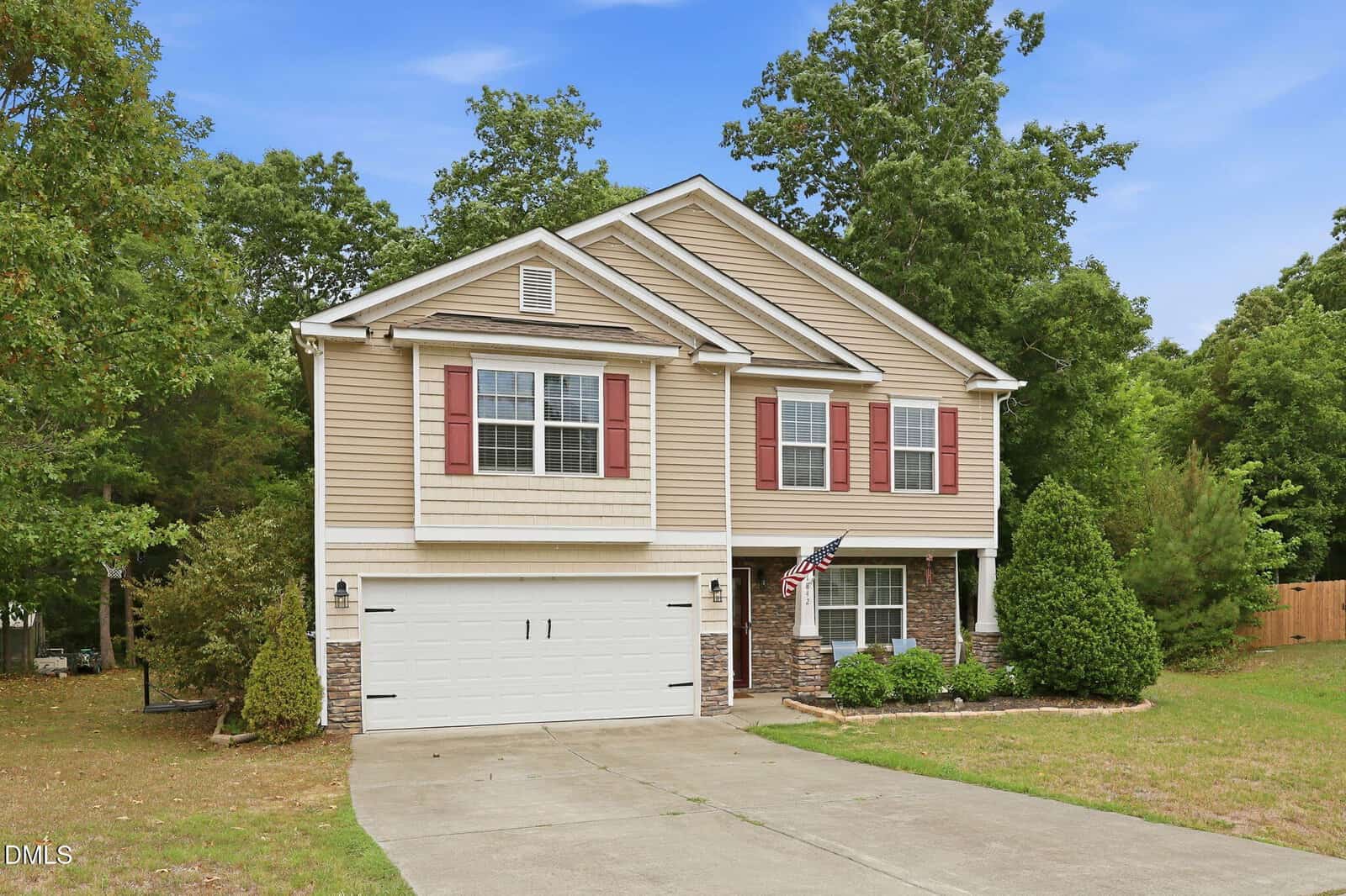 Two-story beige house with red shutters, a white garage door, and stone accents. An American flag hangs near the front door. The house is surrounded by trees and bushes, with a concrete driveway leading up to it.