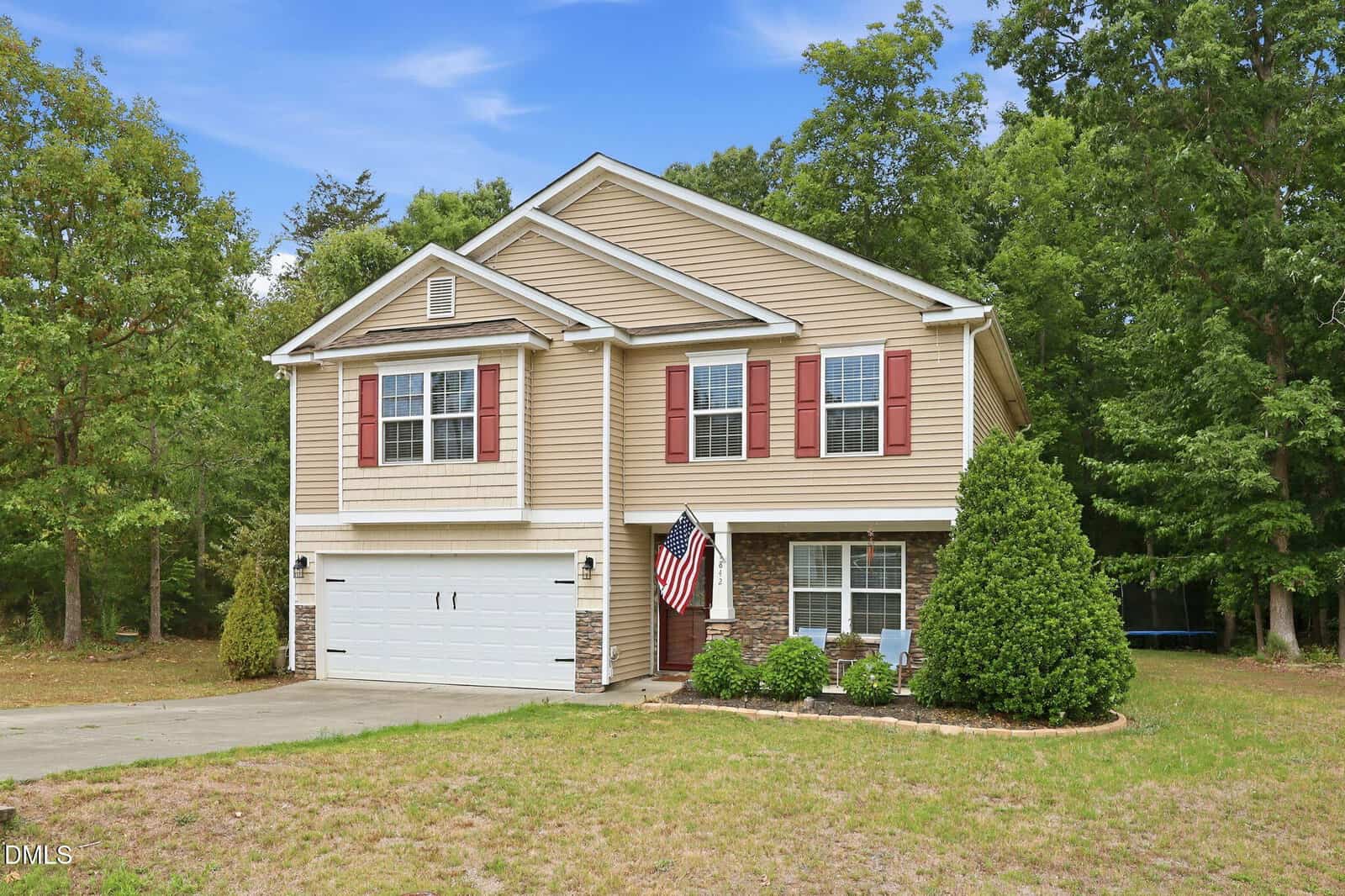A two-story beige house with red shutters at 1842 Fillmore Drive, Creedmoor, featuring a white garage door, an American flag by the entrance, and a driveway, all surrounded by green trees and a grassy yard under a blue sky.