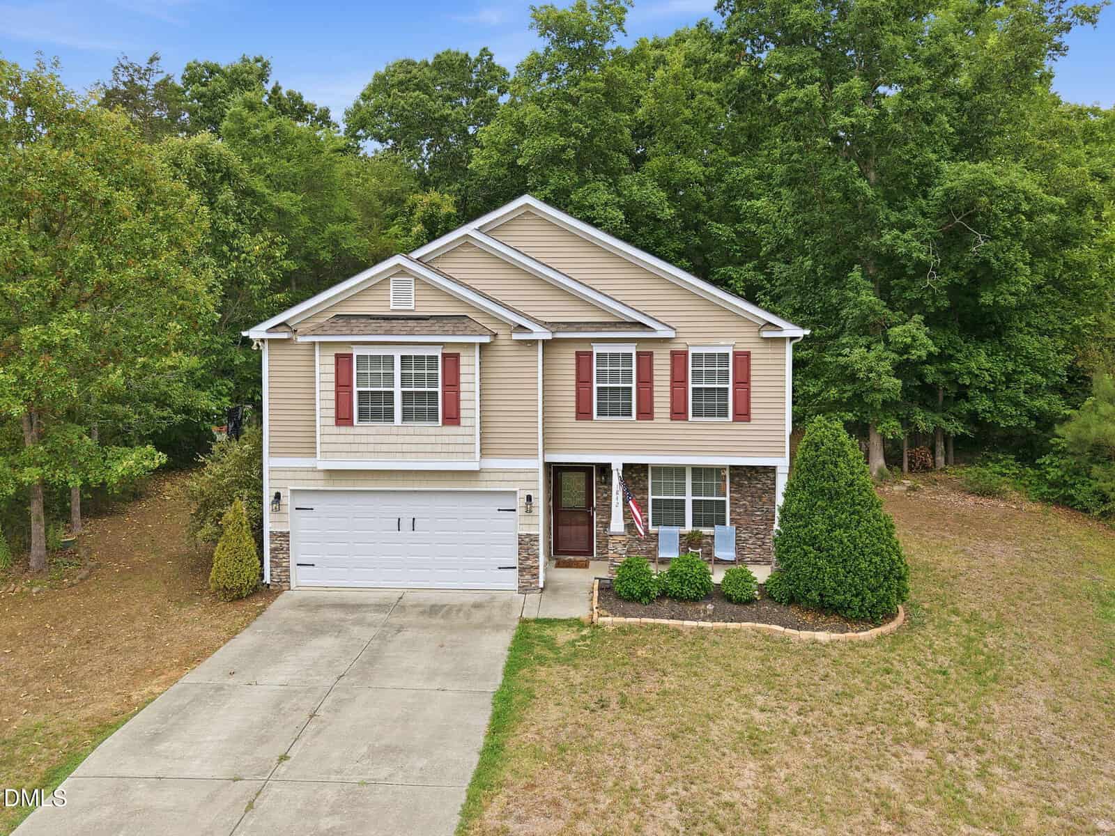 Two-story beige house with red shutters, a double garage, American flag near the entrance, bordered by trees and bushes, with a concrete driveway and patchy grass lawn.