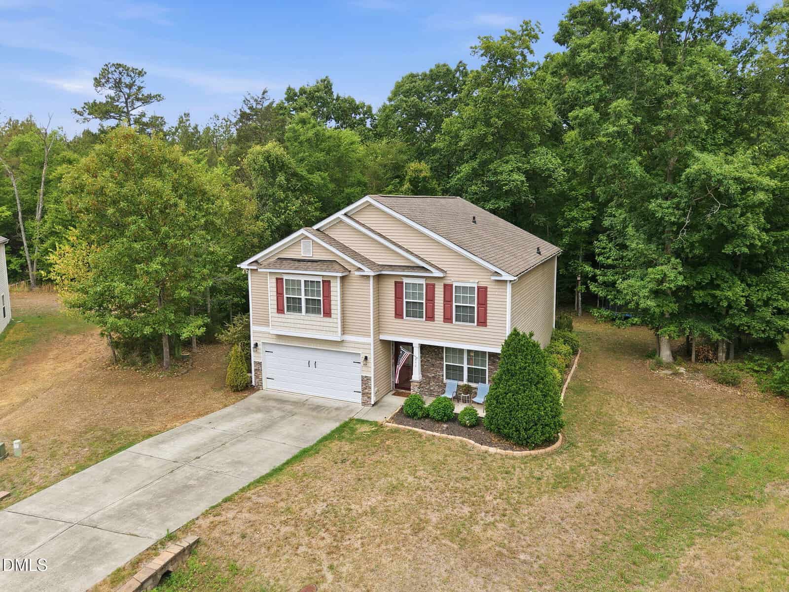Two-story beige house with white trim and red shutters, an attached two-car garage, a concrete driveway, and a small front yard with shrubs. The house is surrounded by trees and has a patchy lawn.