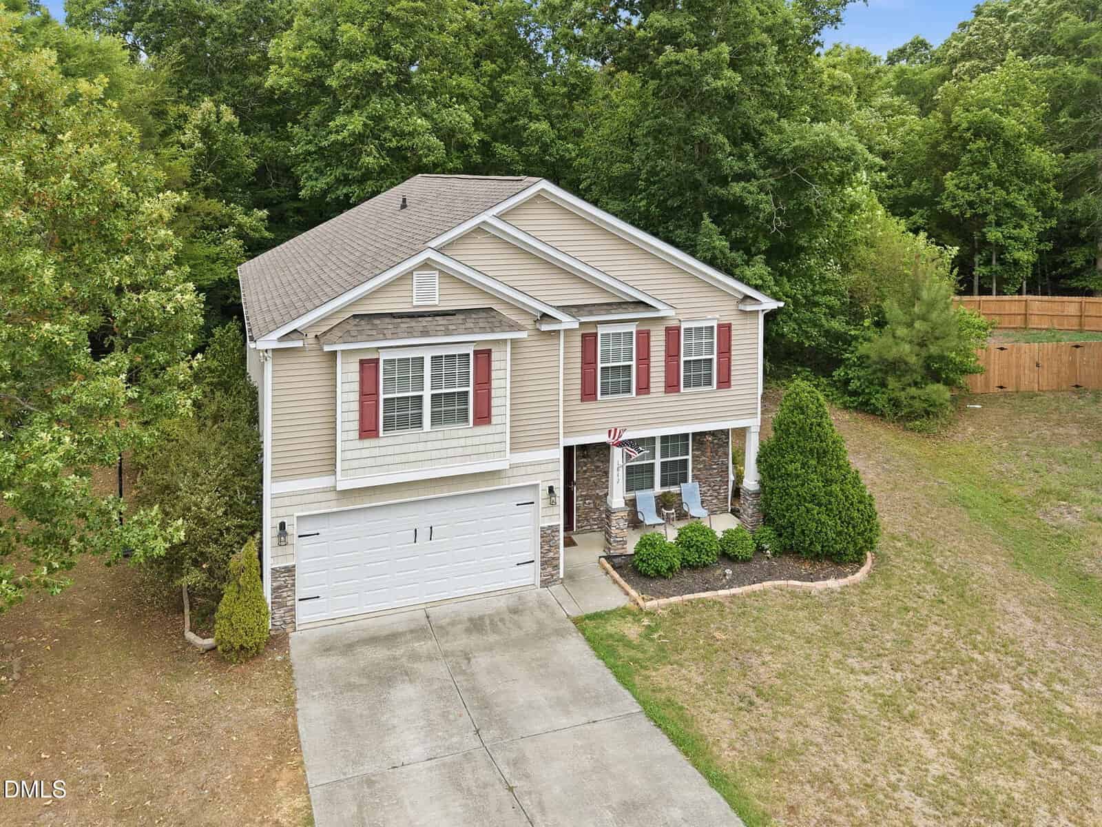 Two-story beige house with red shutters, a double garage, and a concrete driveway. The front yard has shrubs and trees, and the home is bordered by a wooded area and fenced backyard. Two chairs sit on the covered porch.