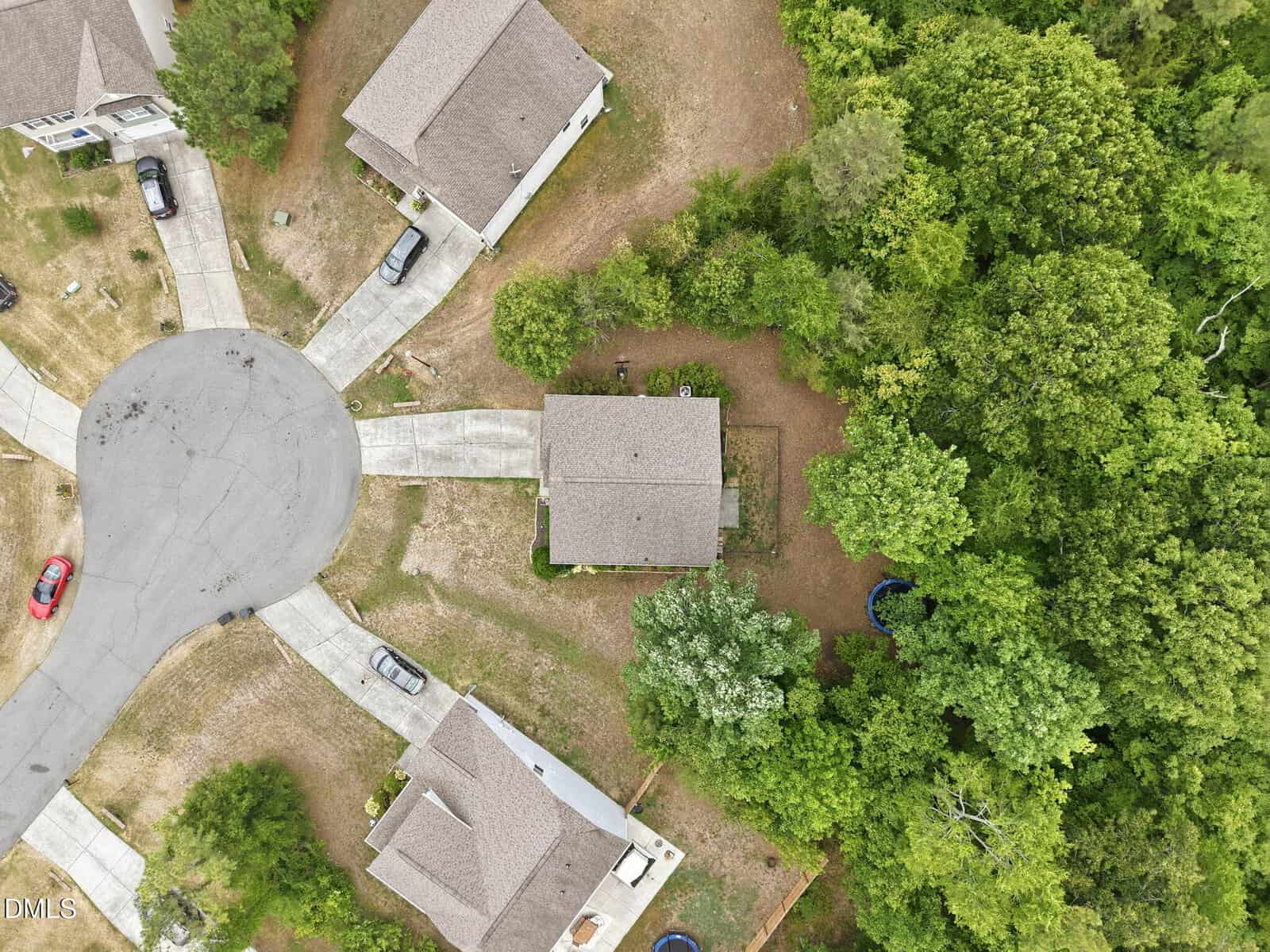 Aerial view of a cul-de-sac with four houses, driveways, cars, and lawns. Dense trees border the right side of the image, and a trampoline is partially visible in a backyard.