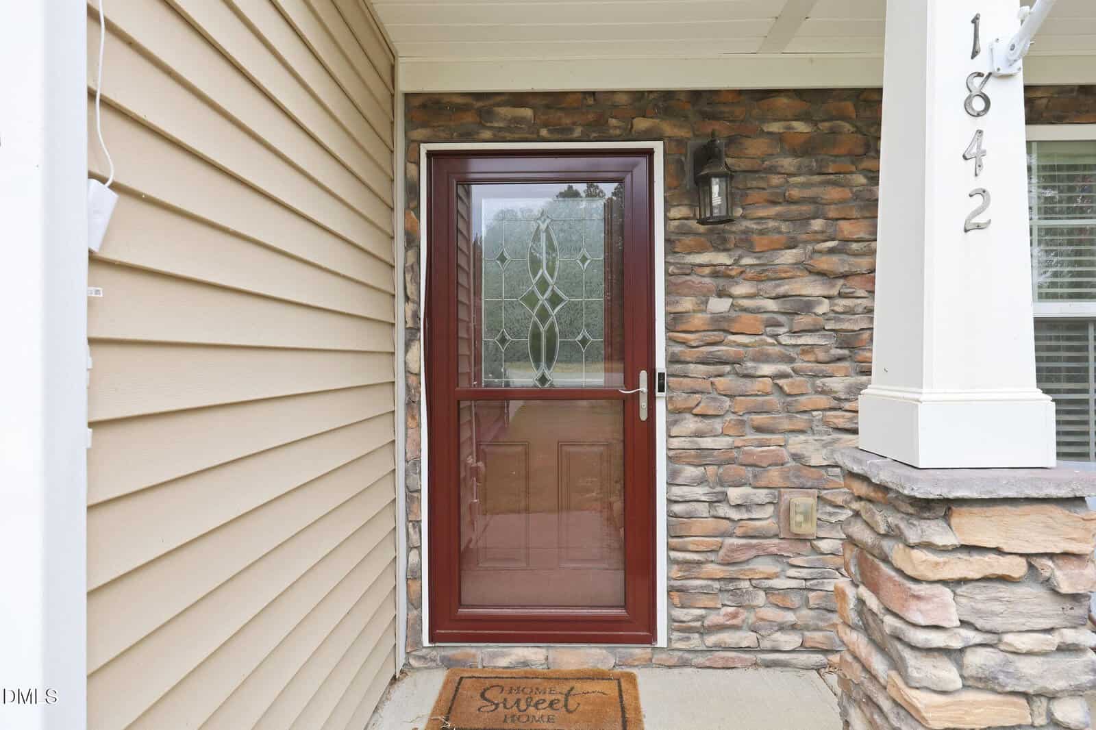 A brown storm door with decorative glass is at the entrance of a house with tan siding and stone accents. The house number 1842 is on a white pillar, and a "Home Sweet Home" doormat lies in front of the door.