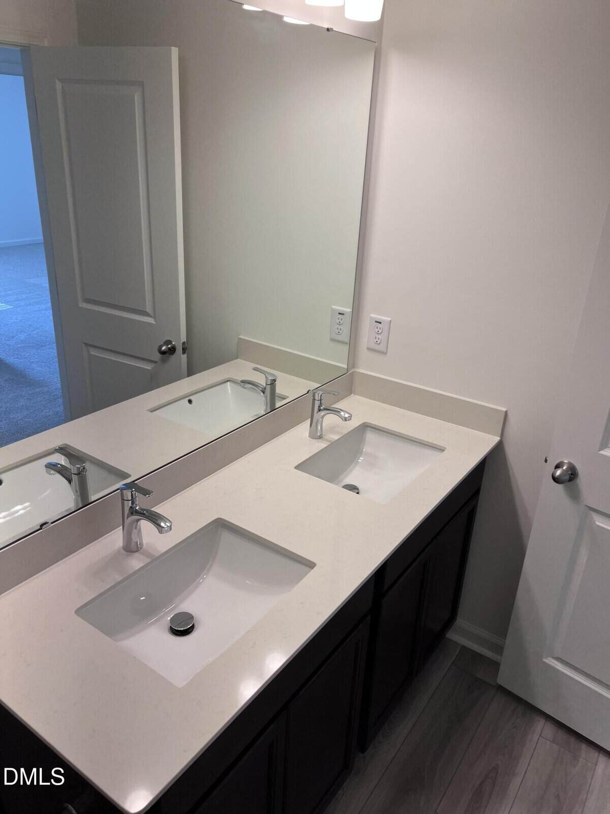 Modern bathroom with a double sink vanity, white countertop, dark cabinets, large mirror, and light gray wood-look flooring. An open door reveals a carpeted room in the background.