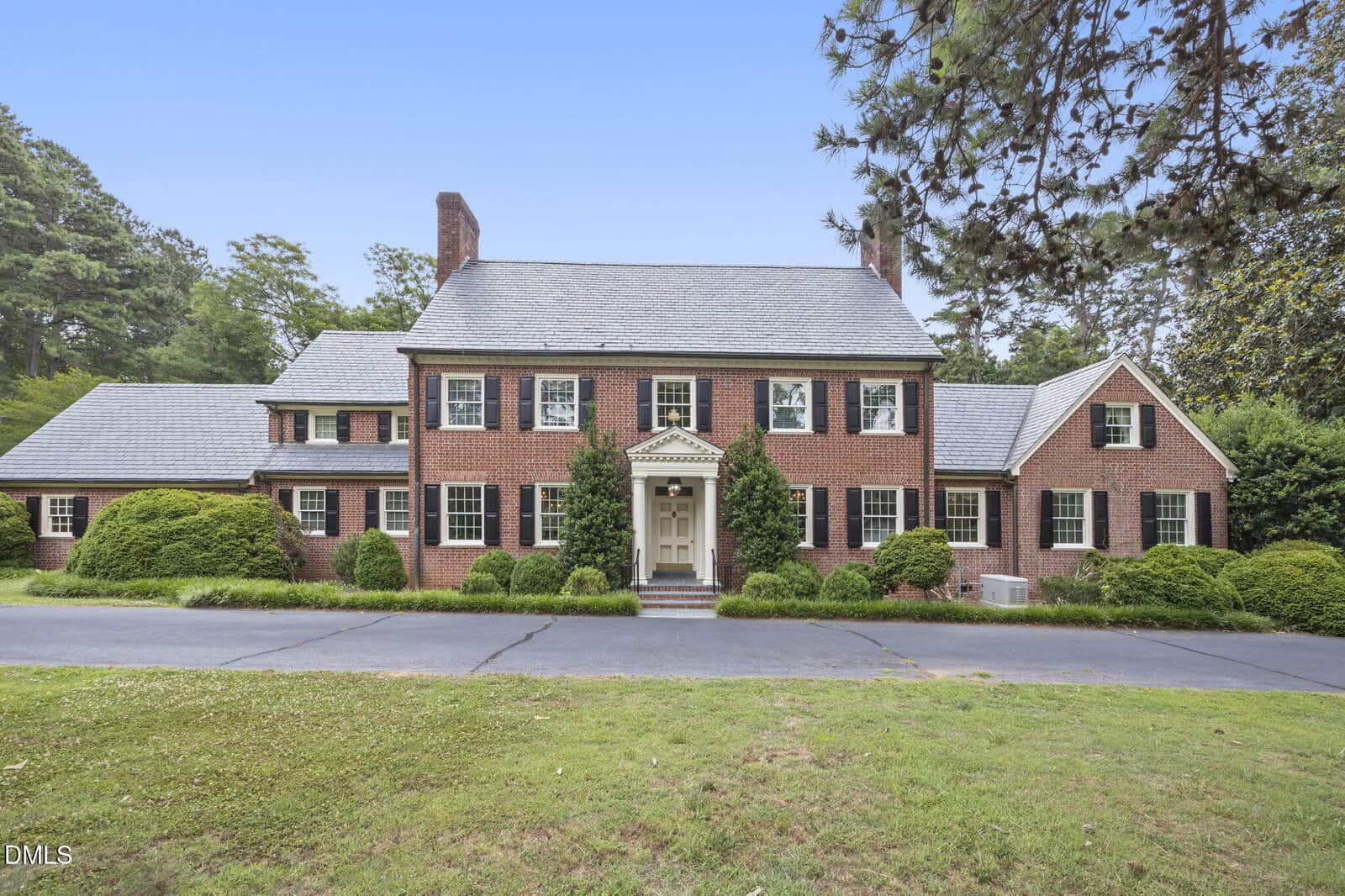 A large two-story brick house with white trim at 1 Lakeside Drive, Henderson, features black shutters, a gray slate roof, and is surrounded by green shrubs and trees, with a paved driveway and grassy front lawn.