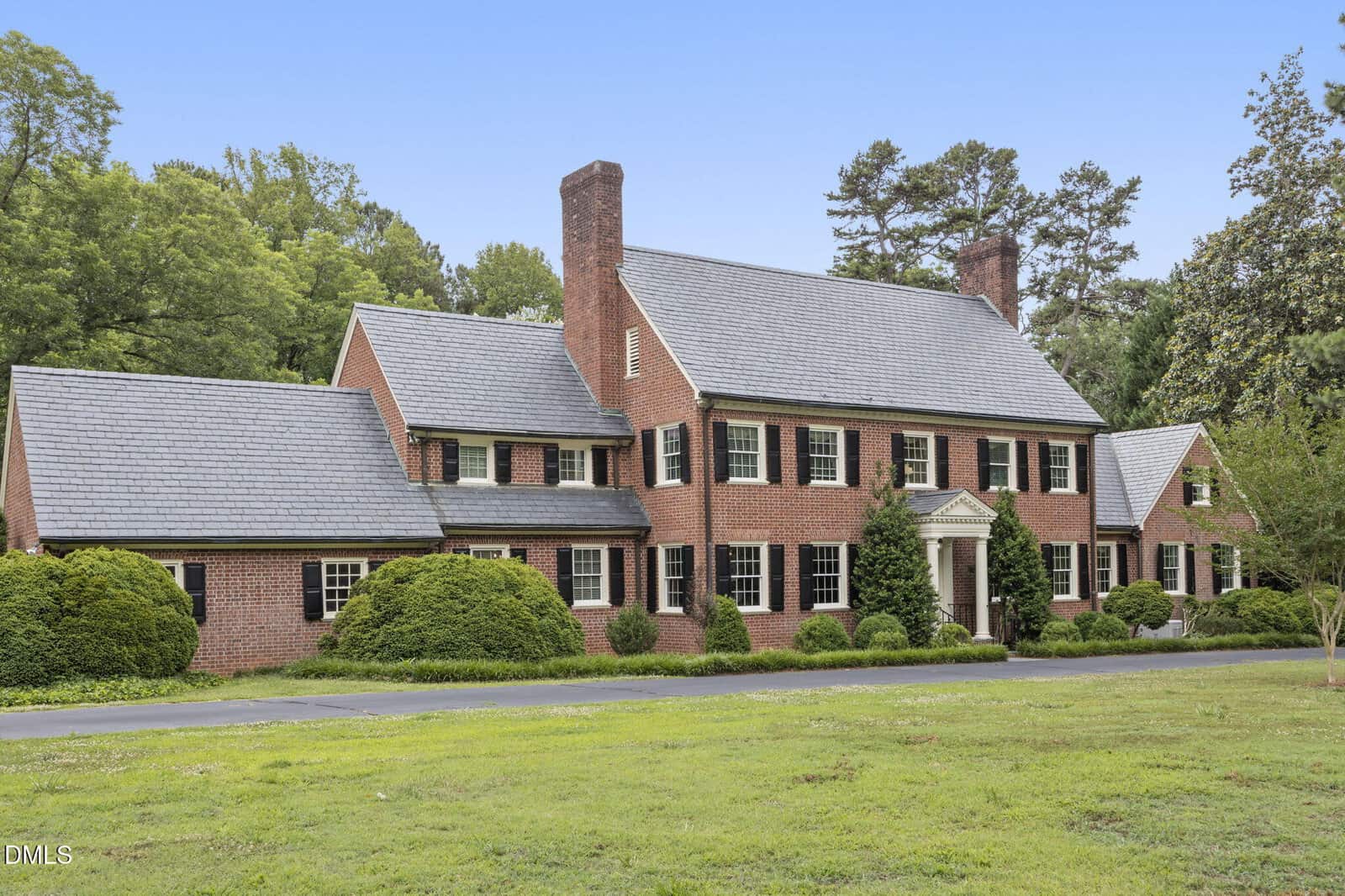 A large two-story red brick colonial-style house with white trim, black shutters, and two chimneys, surrounded by green bushes and trees, with a grassy lawn in the foreground.