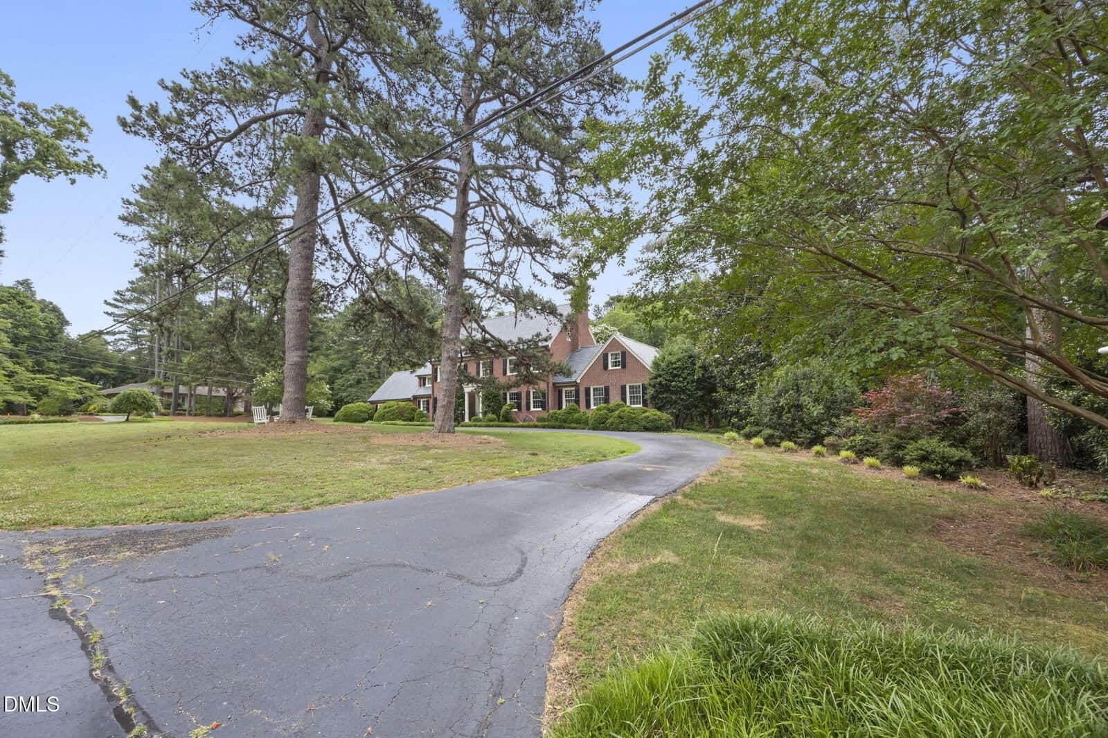 A large brick house with white trim sits behind tall trees, with a curved driveway leading to the entrance, surrounded by a spacious, green lawn and landscaped shrubs.