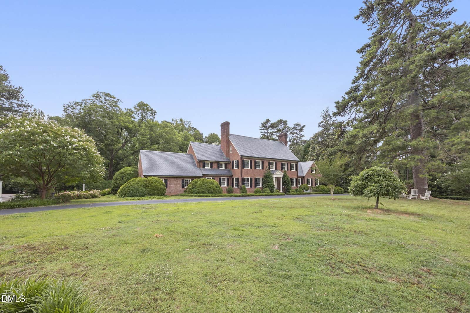 Large brick house with black shutters and a gray roof, surrounded by green lawn, trees, and shrubs on a clear day. A curved driveway leads to the front entrance.