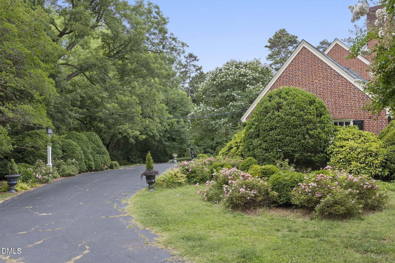A paved driveway curves past a red brick house with manicured bushes, flowering plants, and lush green trees, creating a peaceful, garden-like setting on a clear day.