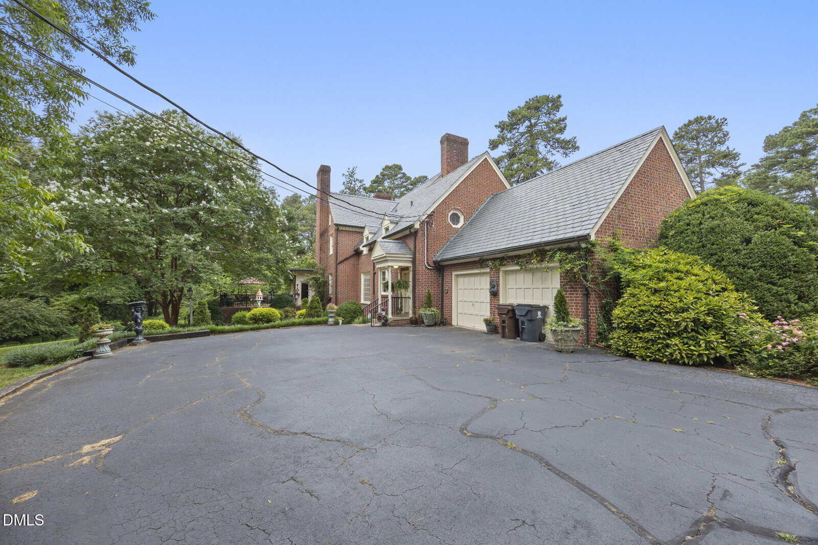 A large brick house with a steep gray roof, two-car garage, and a wide cracked asphalt driveway. Green shrubs and trees surround the property under a clear blue sky. Trash bins are near the garage.