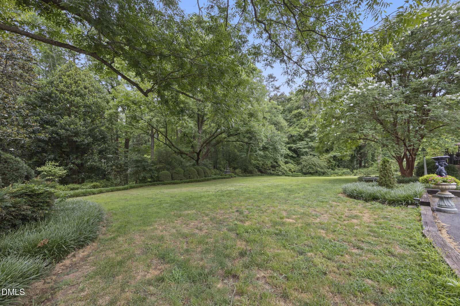 A large, grassy lawn bordered by dense trees and shrubs. A paved path with decorative plants runs along the right side. The sky is partly cloudy, and the area appears well-maintained and lush.
