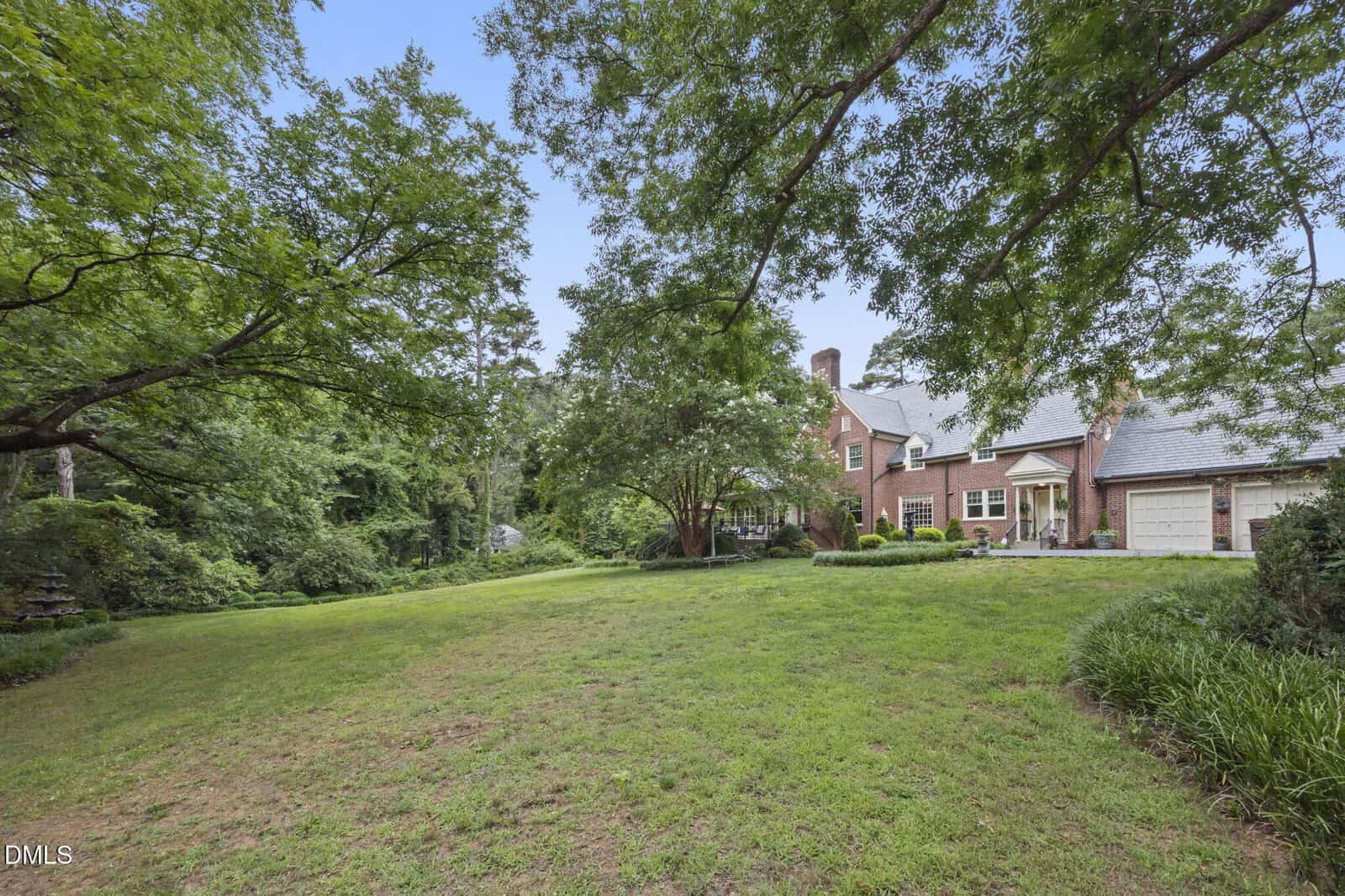 A large brick house with a gray roof sits at the edge of a spacious grassy yard, surrounded by mature trees and lush landscaping on a clear day.