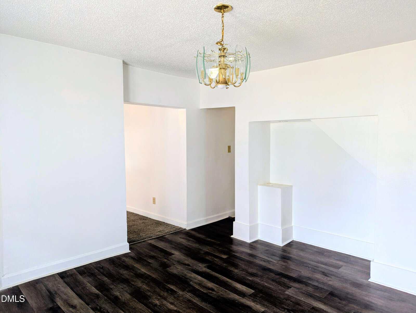Bright room with white walls, dark wood flooring, a brass and glass chandelier, and an open doorway leading to a carpeted area. There is a recessed wall niche next to the doorway.