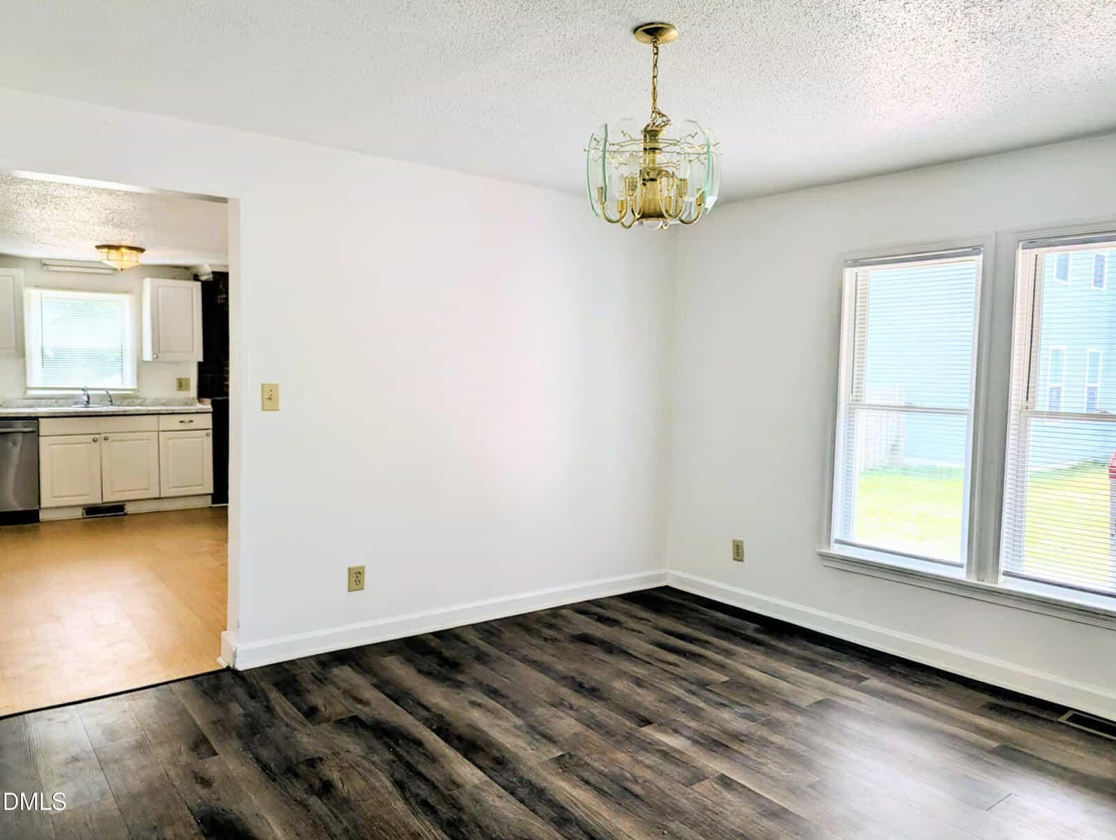 Bright, empty dining room with dark wood flooring, white walls, a brass chandelier, and large windows; kitchen with white cabinets and wood-look floor visible through the doorway.