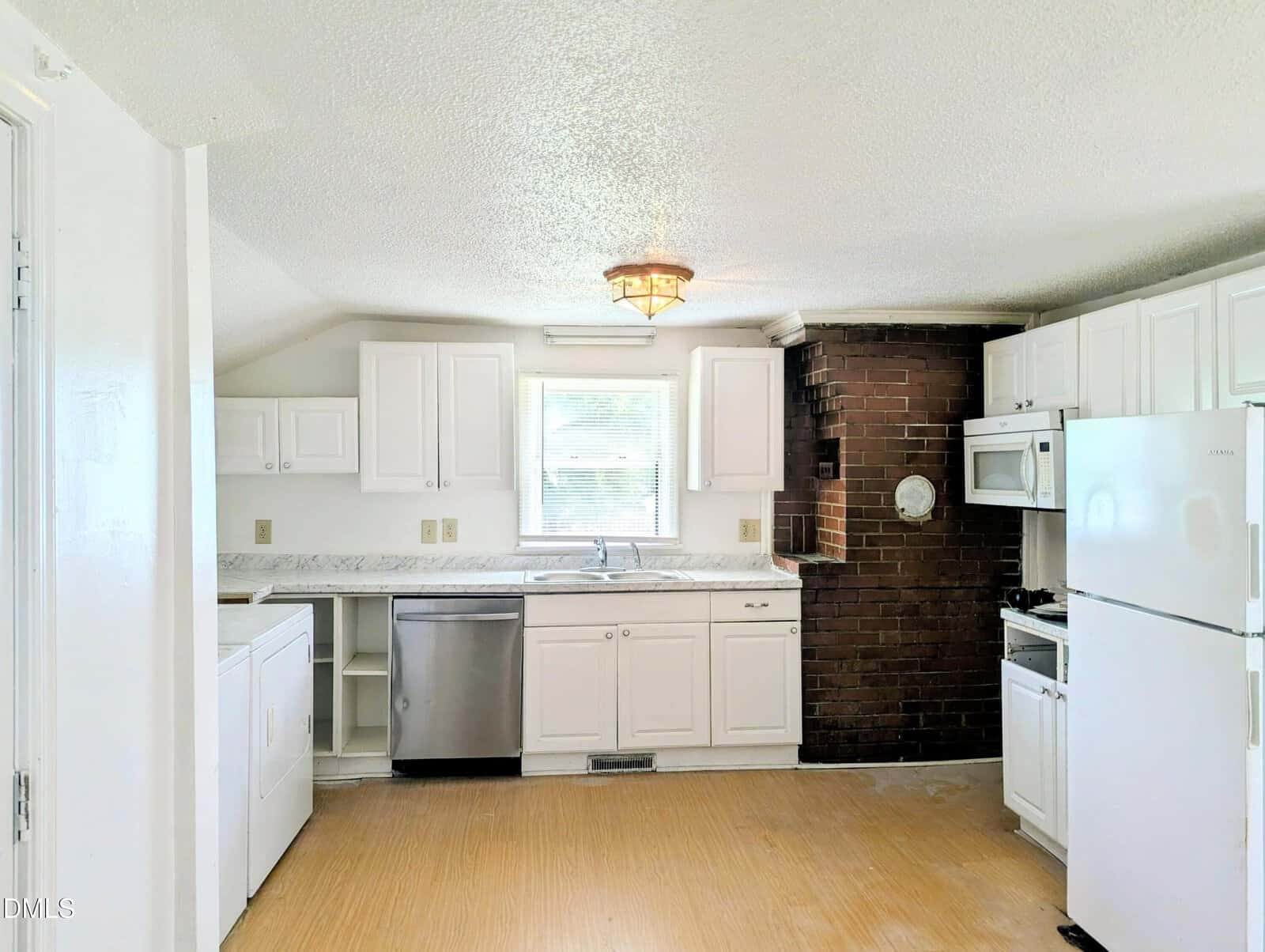 Bright kitchen with white cabinets, stainless steel dishwasher, brick accent wall, white appliances, and a ceiling light. A window above the sink lets in natural light. Light-colored flooring covers the space.