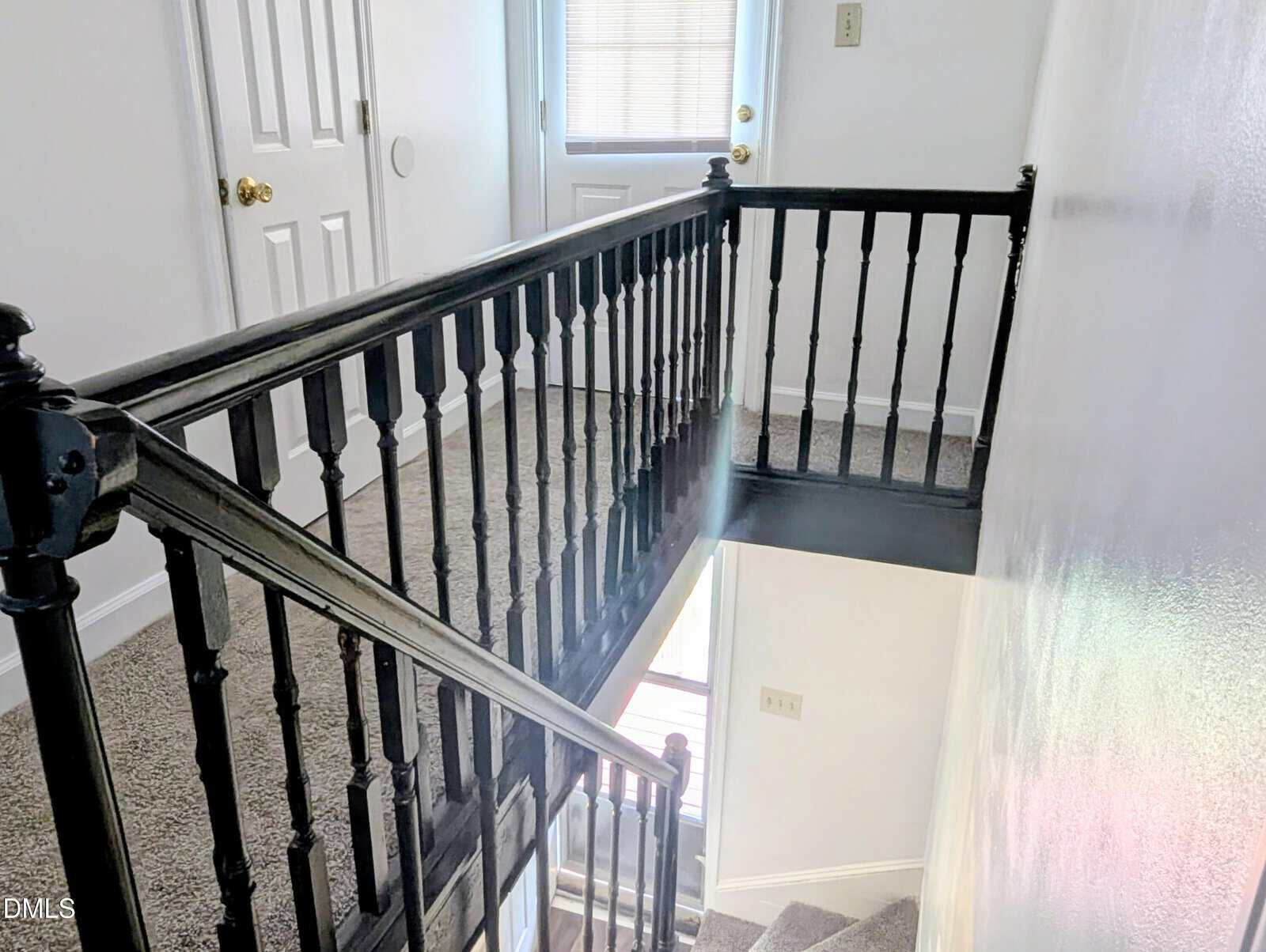 Carpeted staircase with black wooden railing, leading to a small landing with two white doors and a door with blinds. Walls are white and the area is well-lit by natural light.