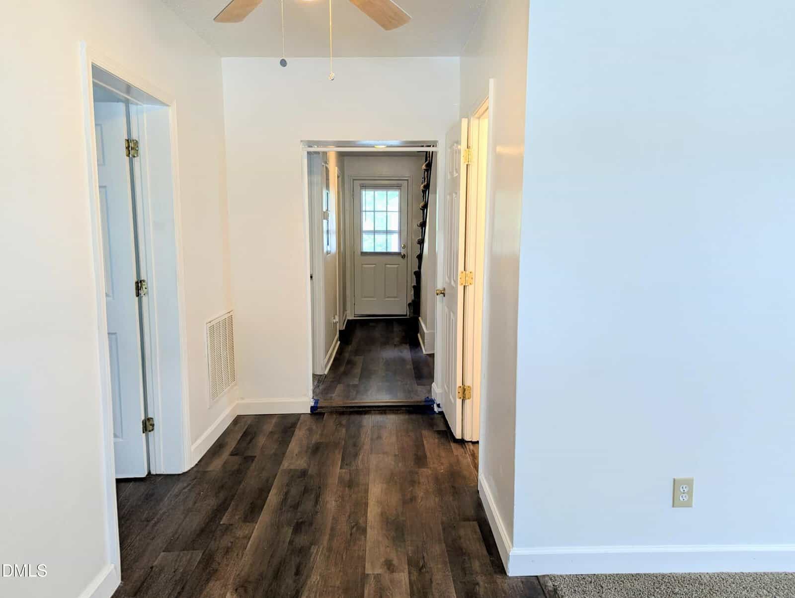 A hallway with dark wood flooring, white walls, and multiple doors. At the end of the hall, there is a white back door with windows and a staircase to the right. A ceiling fan blade is partially visible overhead.
