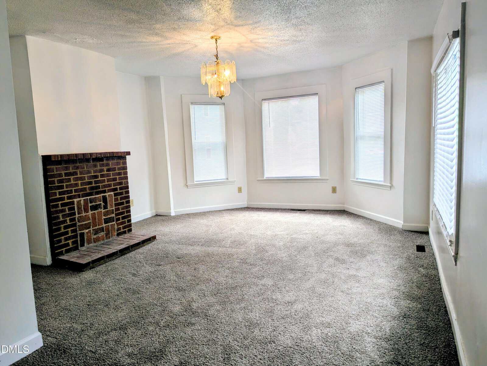 A spacious, carpeted living room with white walls, a brick fireplace on the left, three large bay windows with blinds, and a gold chandelier hanging from the ceiling.