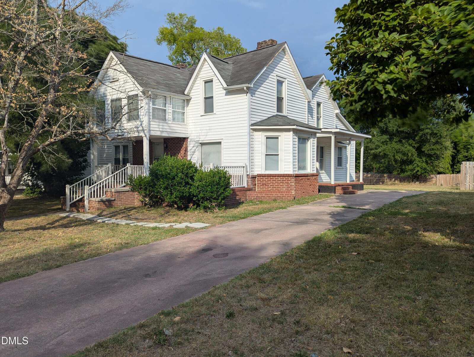 A two-story white house with a gabled roof, brick foundation, and front porch, sits at the end of a long driveway, surrounded by grass, bushes, and trees under a partly cloudy sky.