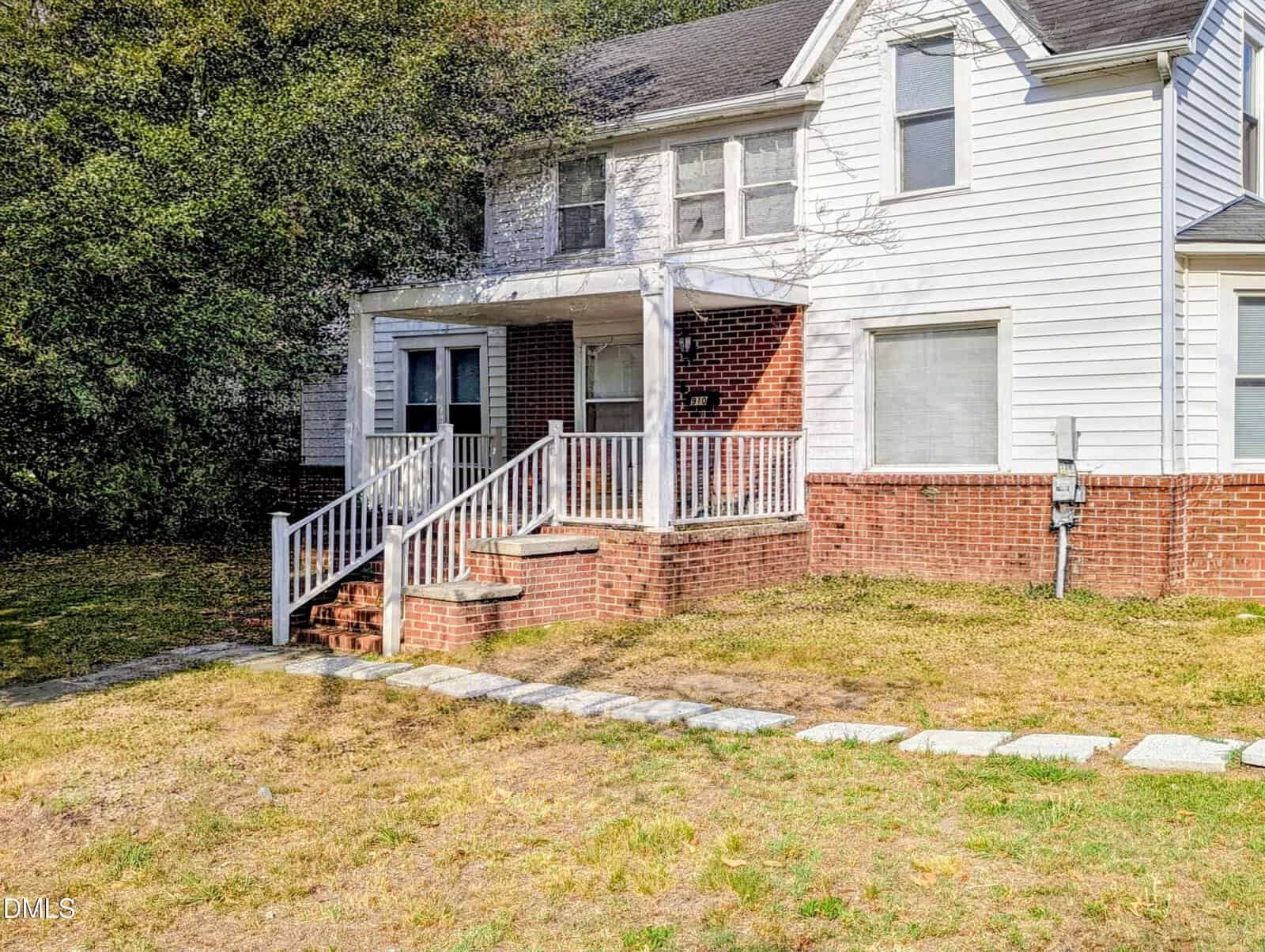 A white two-story house with red brick accents at 910 S Main Street, Louisburg, features a covered front porch and steps to the entrance. The yard offers a stone path, bordered by trees and grass for charming curb appeal.