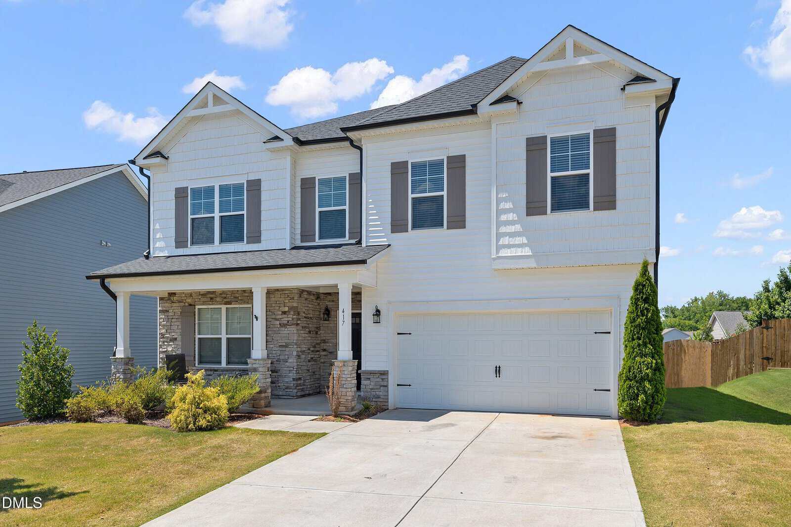 Two-story suburban house with white siding, grey shutters, a covered front porch with brick accents, and a double garage. There is a green lawn, shrubs, and a clear blue sky with scattered clouds.