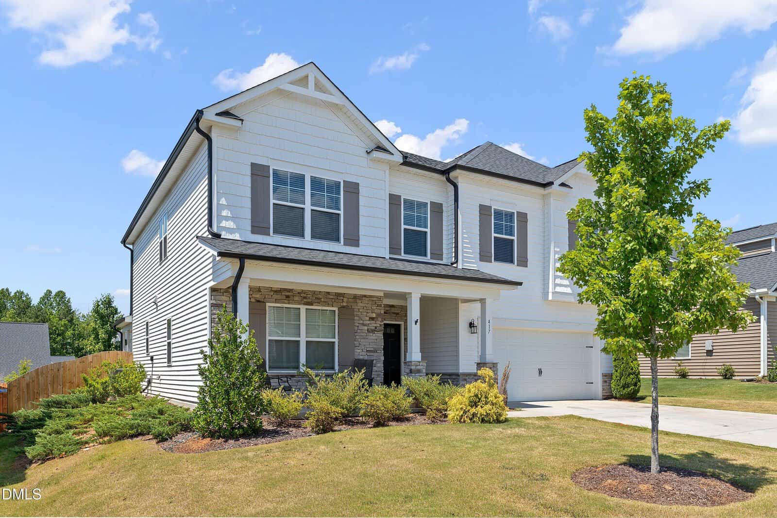 Two-story modern suburban house with white siding, gray shingles, black shutters, and a covered front porch. The front yard has a small tree, shrubs, and a concrete driveway leading to a double garage.