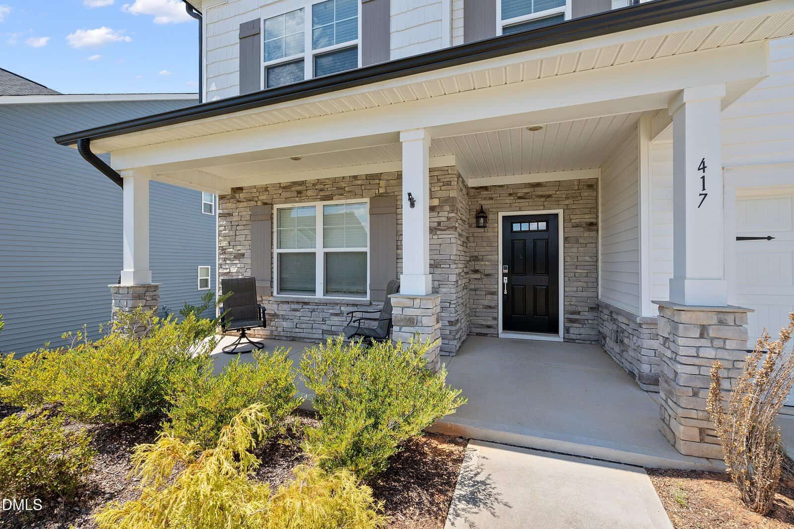 A modern house with a covered front porch, stone accents, black front door, two chairs, and shrubs in the landscaped yard. The house number 417 is visible on a column near the entrance.