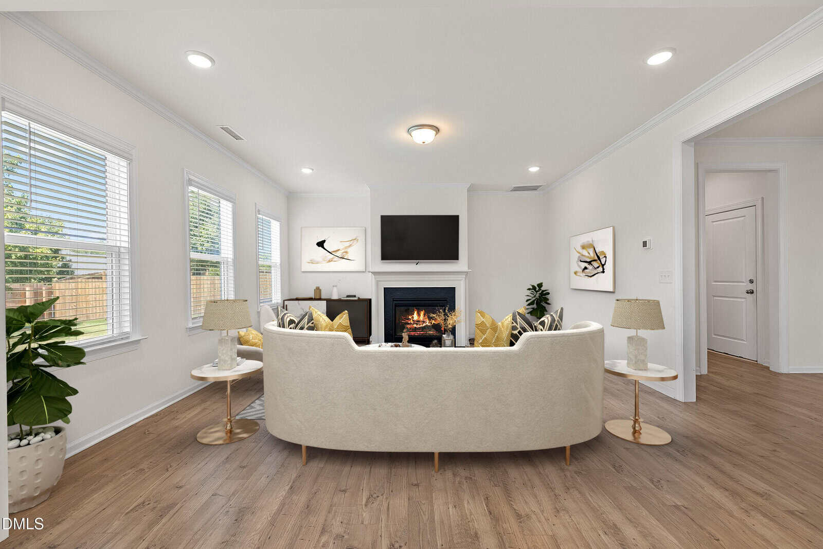 A bright living room with a cream sofa, gold-accented tables and lamps, a fireplace, wall art, a flat-screen TV above the mantel, large windows, wood floors, and a potted plant in the corner.
