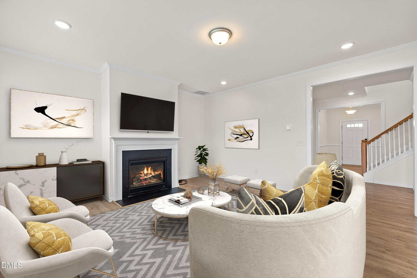 A cozy living room with a fireplace, wall-mounted TV, modern art, light-colored furniture, a round coffee table, and decorative pillows. Wooden floors and a staircase are visible in the background.
