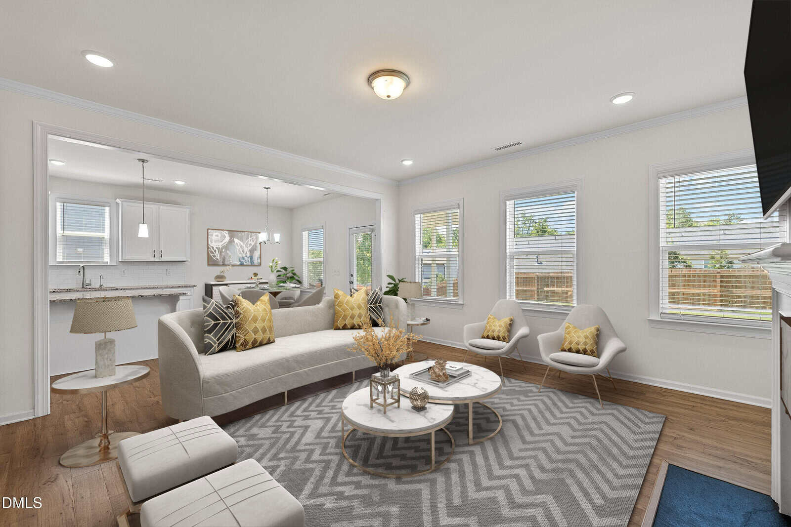 Bright living room with beige sofa, modern chairs, round coffee tables, patterned rug, and open view to a white kitchen. Large windows let in natural light; decorative pillows and plants add warmth.