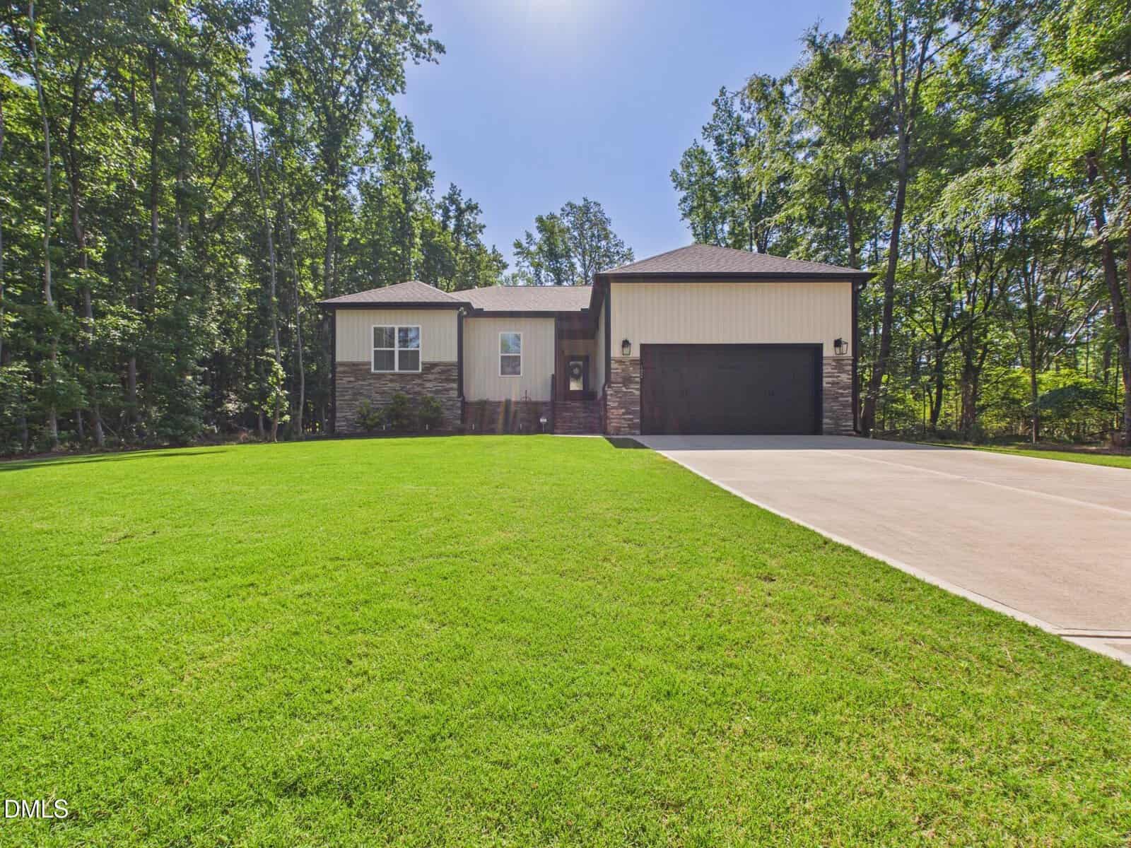A modern single-story house at 132 Mohawk Drive, Louisburg, with a stone and siding exterior, black garage door, and a wide driveway, surrounded by a large, neatly mowed lawn and tall trees under a clear blue sky.