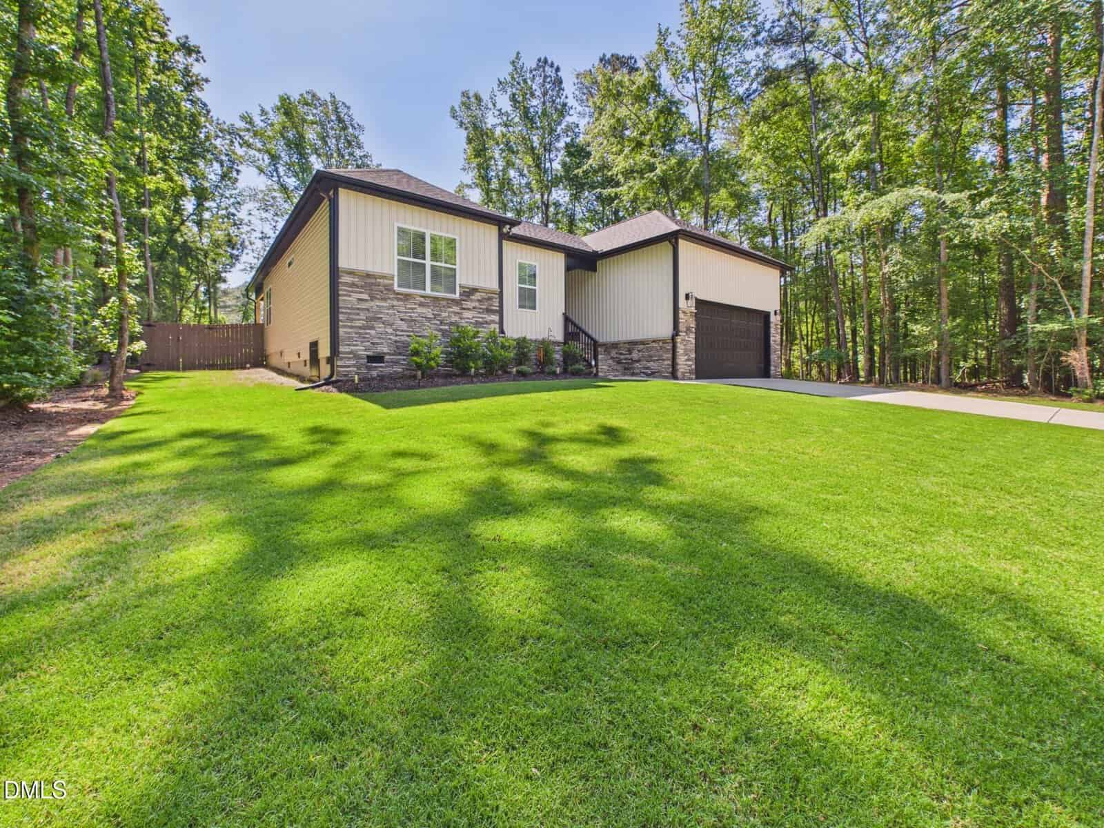 Single-story house with beige siding and stone accents, a dark-colored roof, and a double garage. The house is surrounded by lush green lawn and tall trees under a clear blue sky.