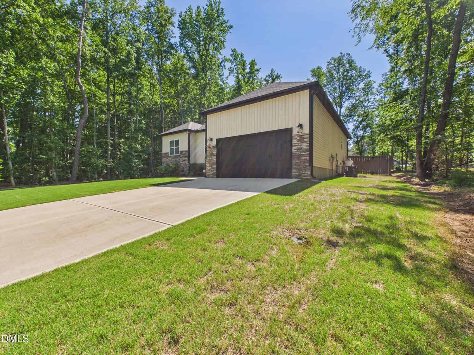 A modern house with beige siding and stone accents, a large dark brown garage door, and a wide concrete driveway, surrounded by green grass and tall trees on a sunny day.