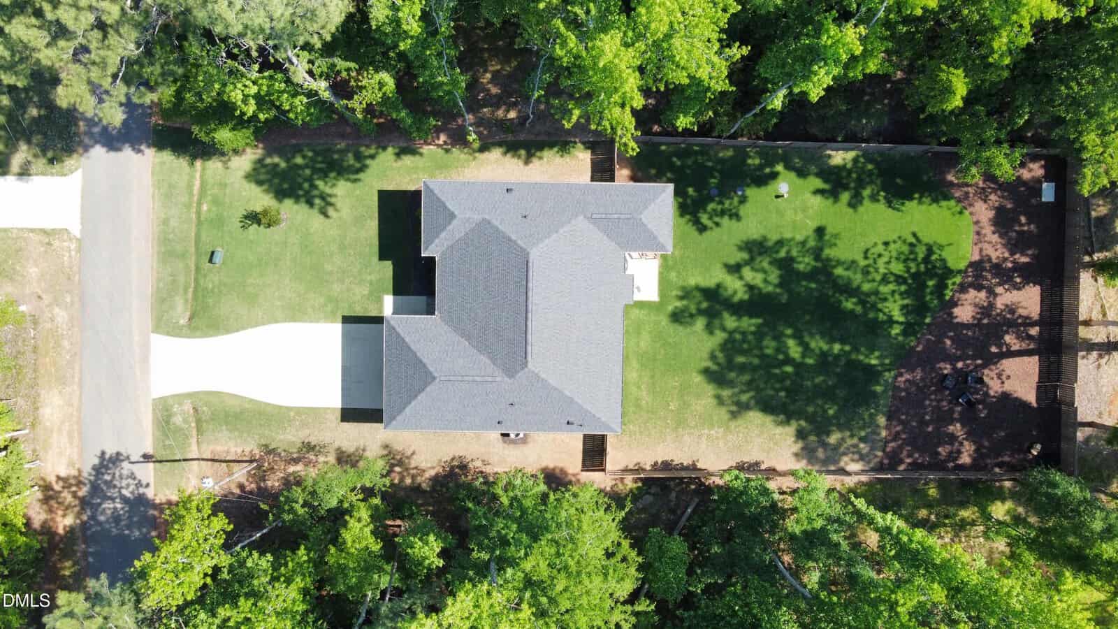 Aerial view of a single-story house with a dark gray roof, white driveway, and a large green lawn, surrounded by trees casting shadows across the yard.