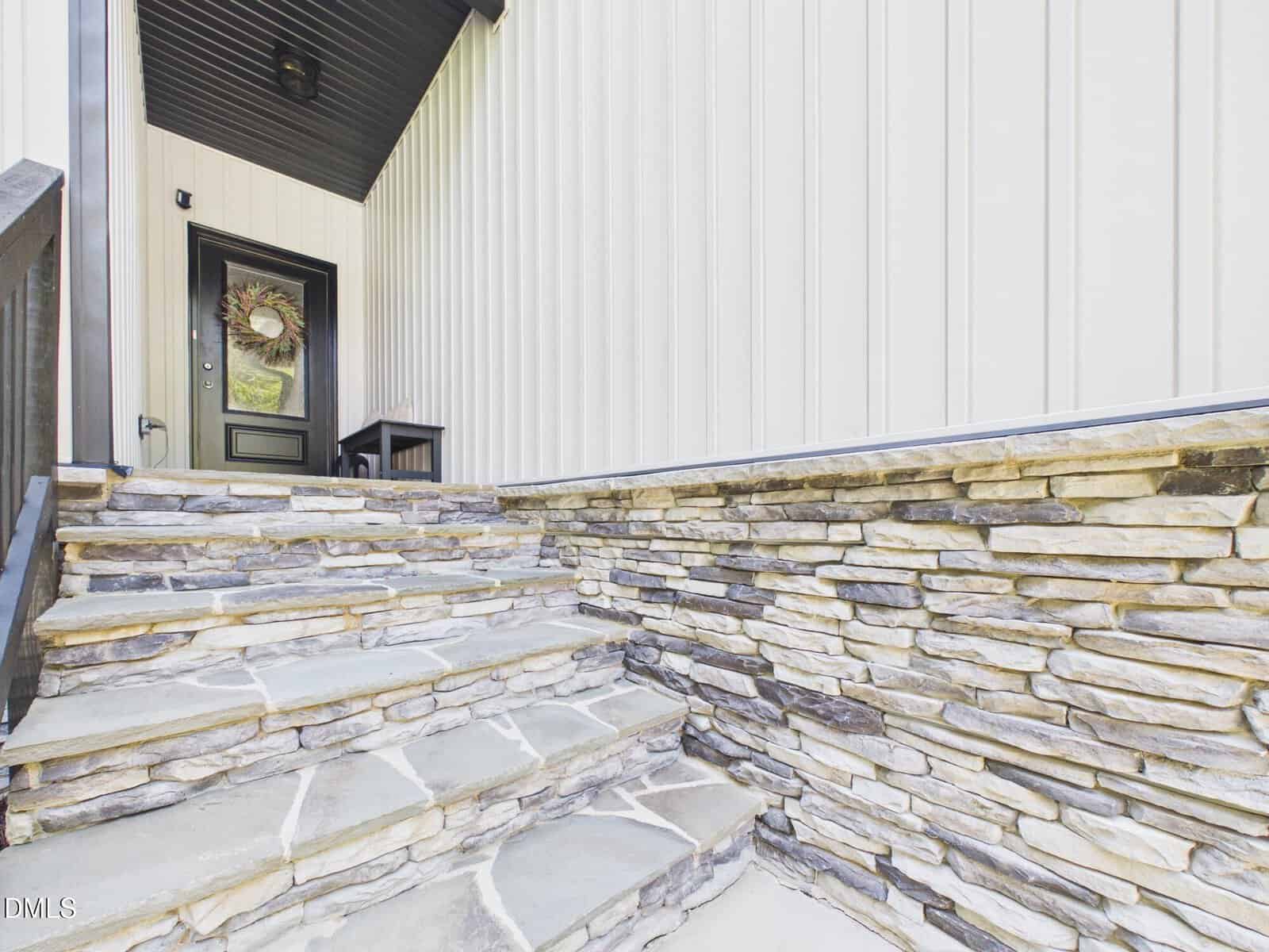 Stone steps lead to a black door with a wreath. The entryway has beige vertical siding and stacked stone walls. A small black bench sits near the door.