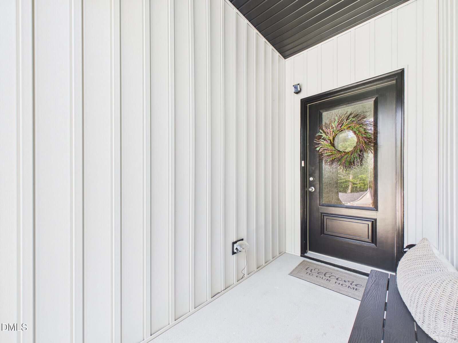A modern entryway with vertical white panel walls, a black door featuring a wreath, a welcome mat, and part of a bench visible in the corner.