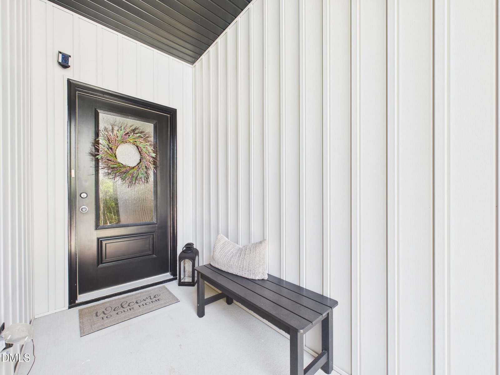 A black front door with a wreath, a “Welcome to our home” doormat, a black bench with a pillow, a lantern, and white vertical siding on the porch.