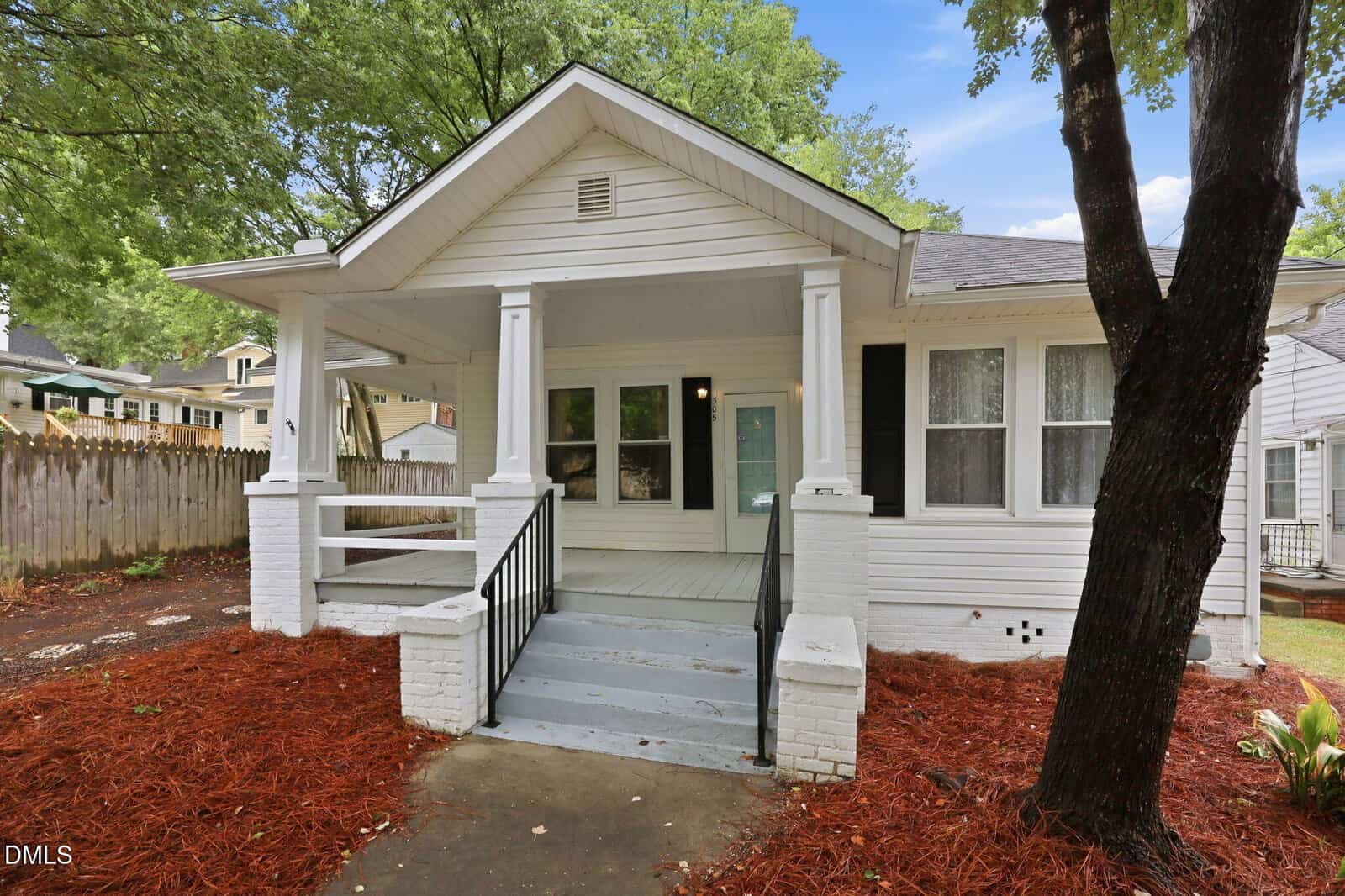 Charming white single-story home at 305 Gilliam Street, Oxford, with a covered front porch, columns, black shutters, and a blue door. Enjoy landscaped yards with red mulch, trees, and a wooden fence on the left side.
