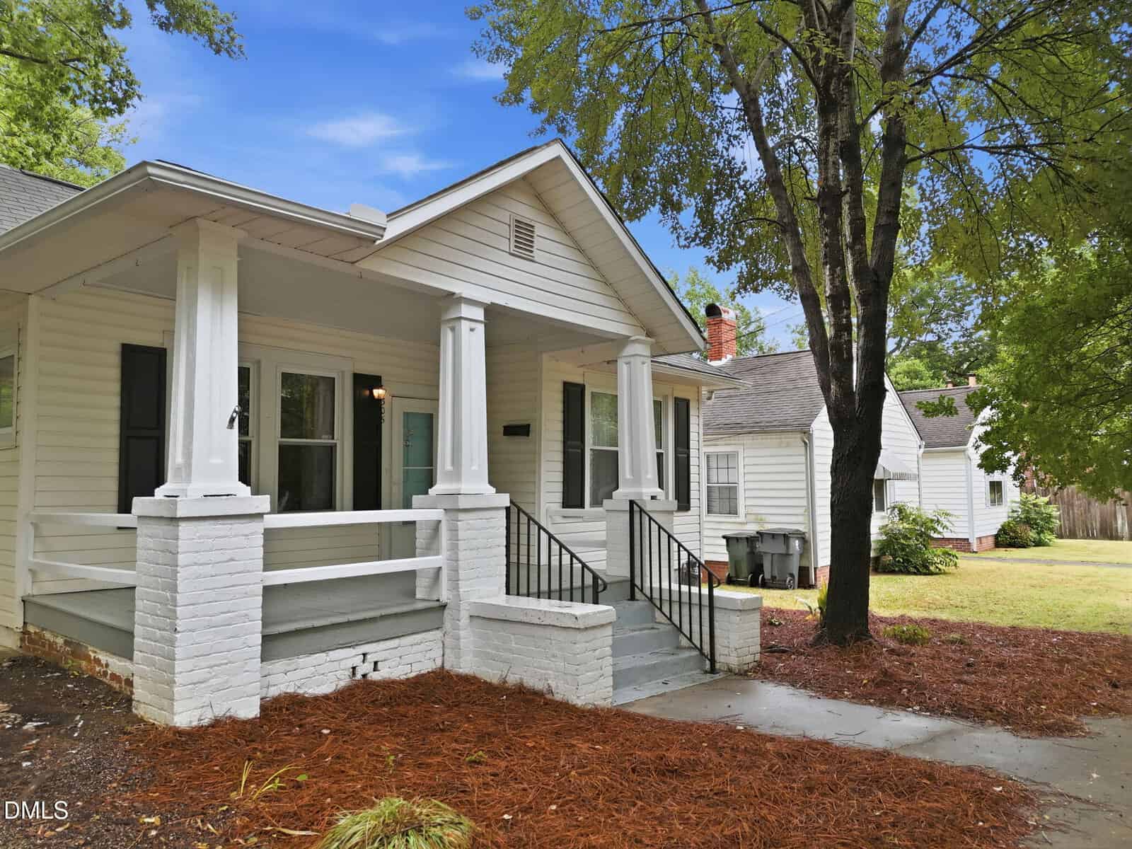 A white single-story house with a covered front porch, black railings, and columns, surrounded by trees and a grassy yard under a blue sky.