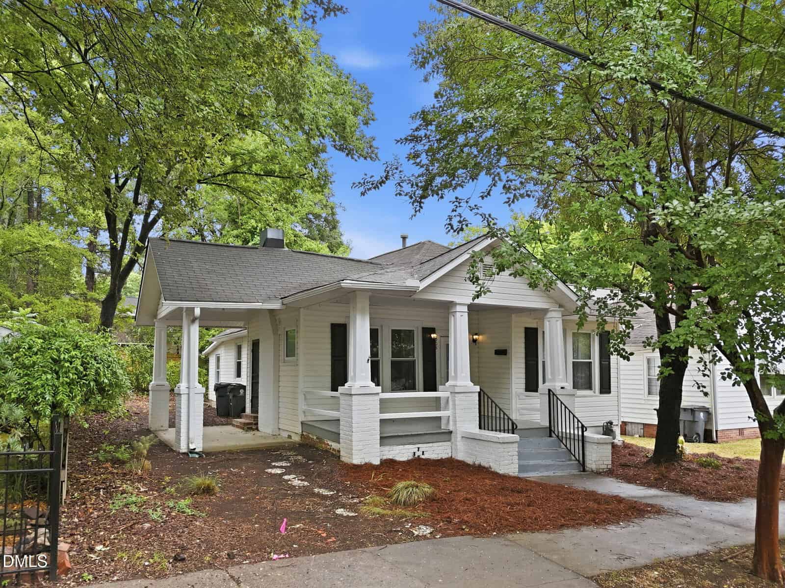 A charming single-story white house with a covered front porch, brick columns, a carport, and black railings, surrounded by trees and greenery under a bright blue sky.