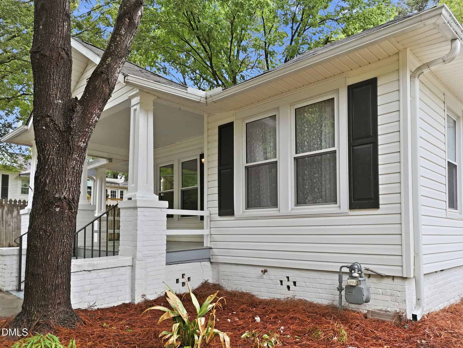 A white house with black shutters, a covered front porch, and large windows. Mulch and small plants are in the landscaped area beside the house, and a tree stands in the foreground under a partly cloudy sky.