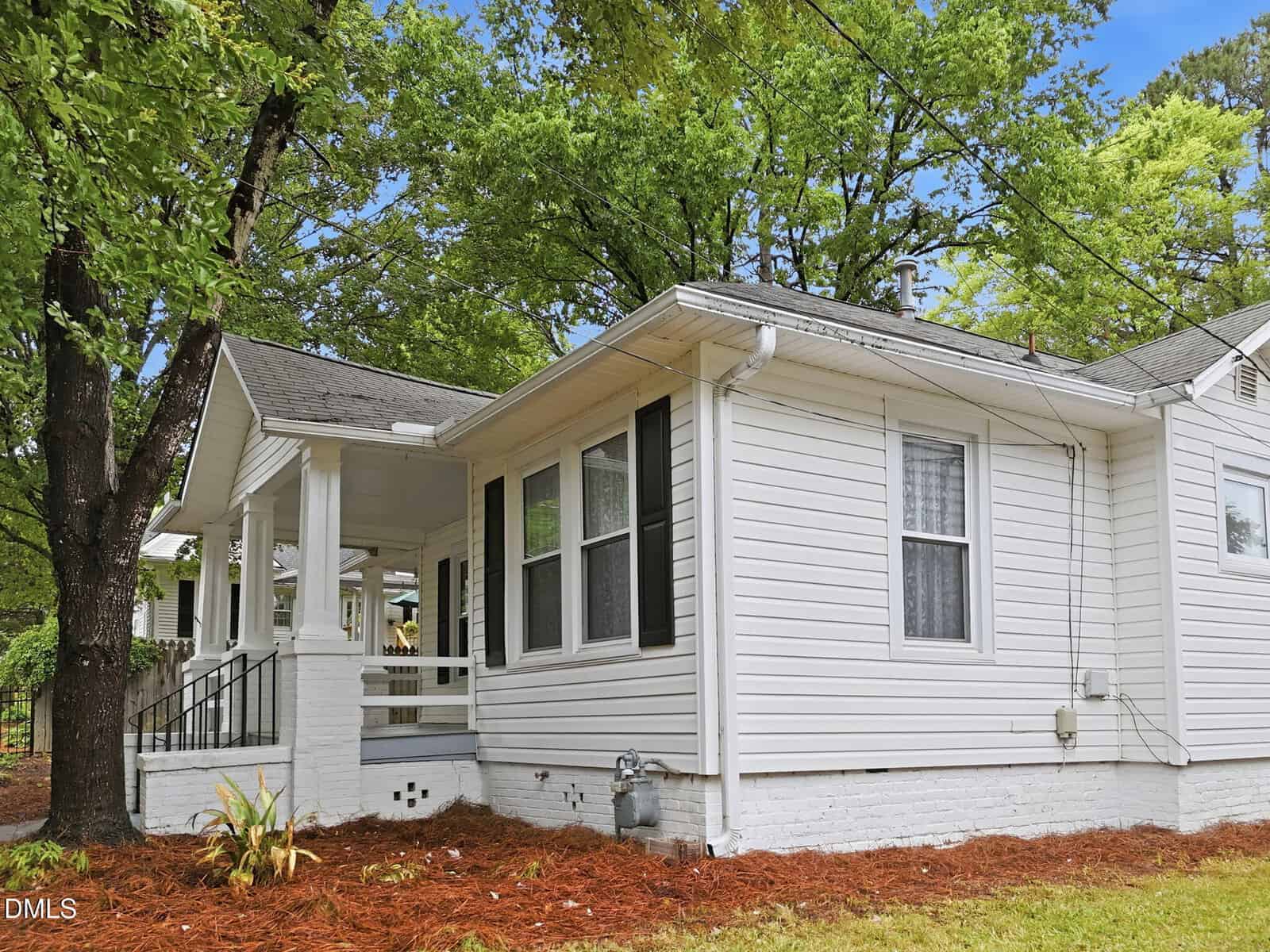 White single-story house with black shutters, large windows, and a covered front porch. Surrounded by green trees and a yard with mulch and plants. A bright, clear day.