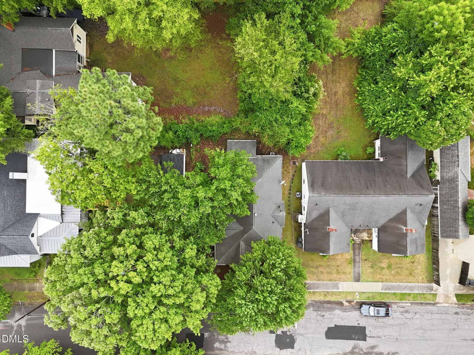 Aerial view of three houses surrounded by green trees, with a grassy yard between them. One car is parked on the street in front of the houses.