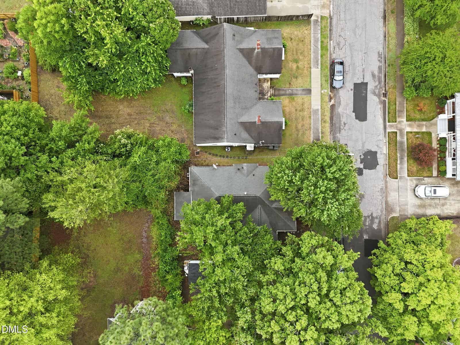 Aerial view of two houses surrounded by trees, with driveways leading to a street where two cars are parked. The roofs are partially obscured by the dense green foliage.
