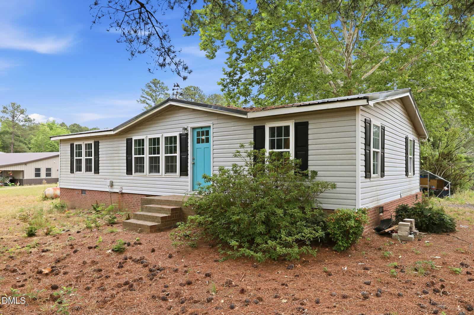 Single-story house with light gray siding, white trim, and black shutters. A bright turquoise front door sits at the top of brick steps. The yard is covered in pine needles with scattered bushes and trees in the background.