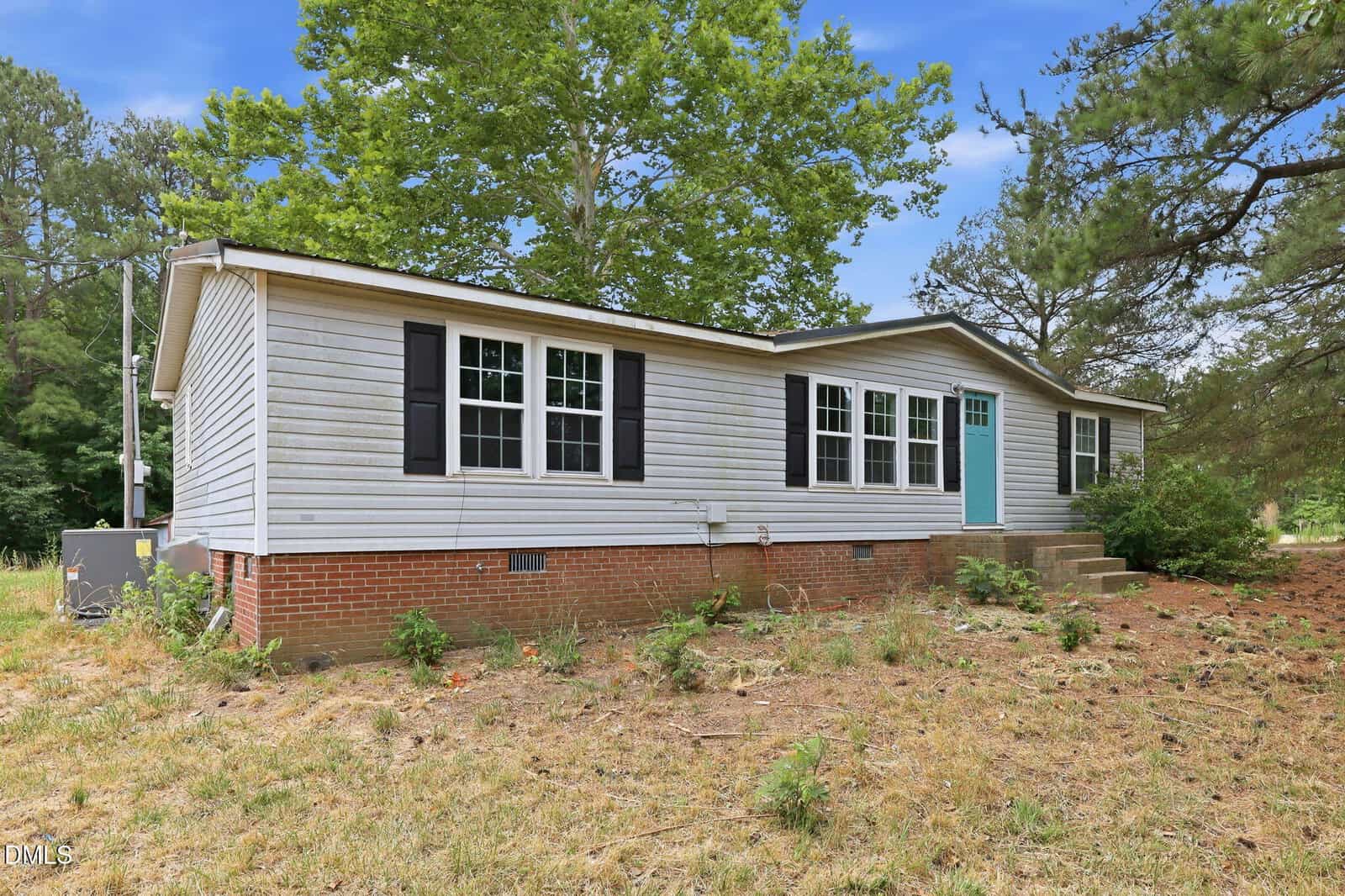 Single-story house with light gray siding, black shutters, and a teal front door, set on a brick foundation. The yard is mostly dry grass with some patches of greenery and tall trees in the background.