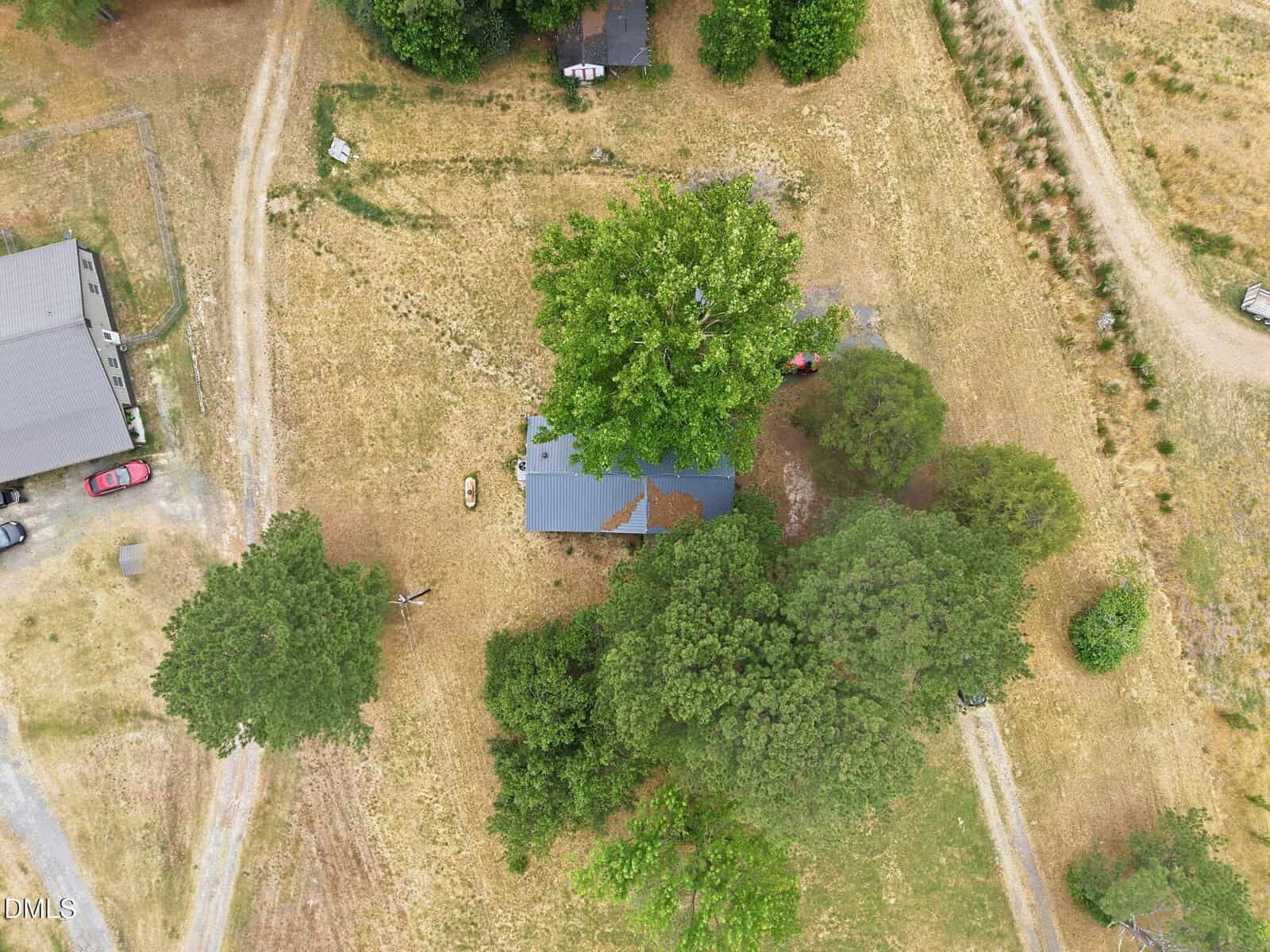 Aerial view of a rural property with a house partially shaded by trees, surrounded by dry grass, a dirt driveway, a few vehicles, and some scattered trees and shrubs.
